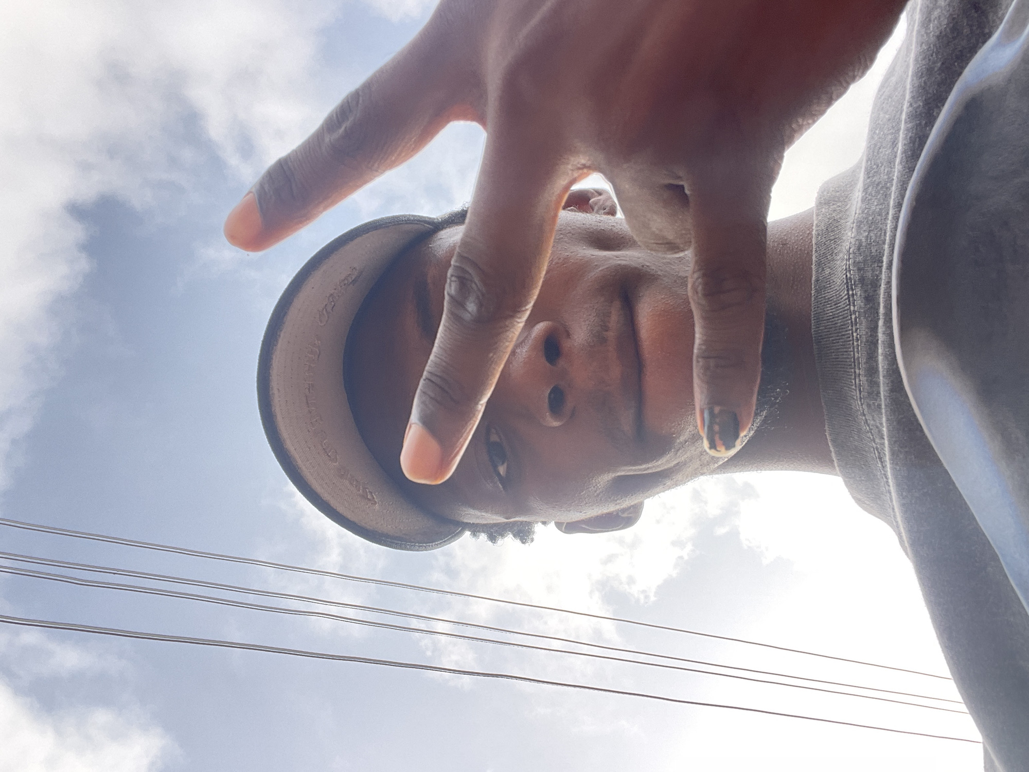 A low-angle shot depicts a man looking down at the camera. He is wearing a baseball cap. His hand is held up, fingers spread, partially obscuring his face. Power lines are visible in the background, against a sky with scattered clouds. The location, Lokoja, Nigeria, is not discernible within the image without further information.