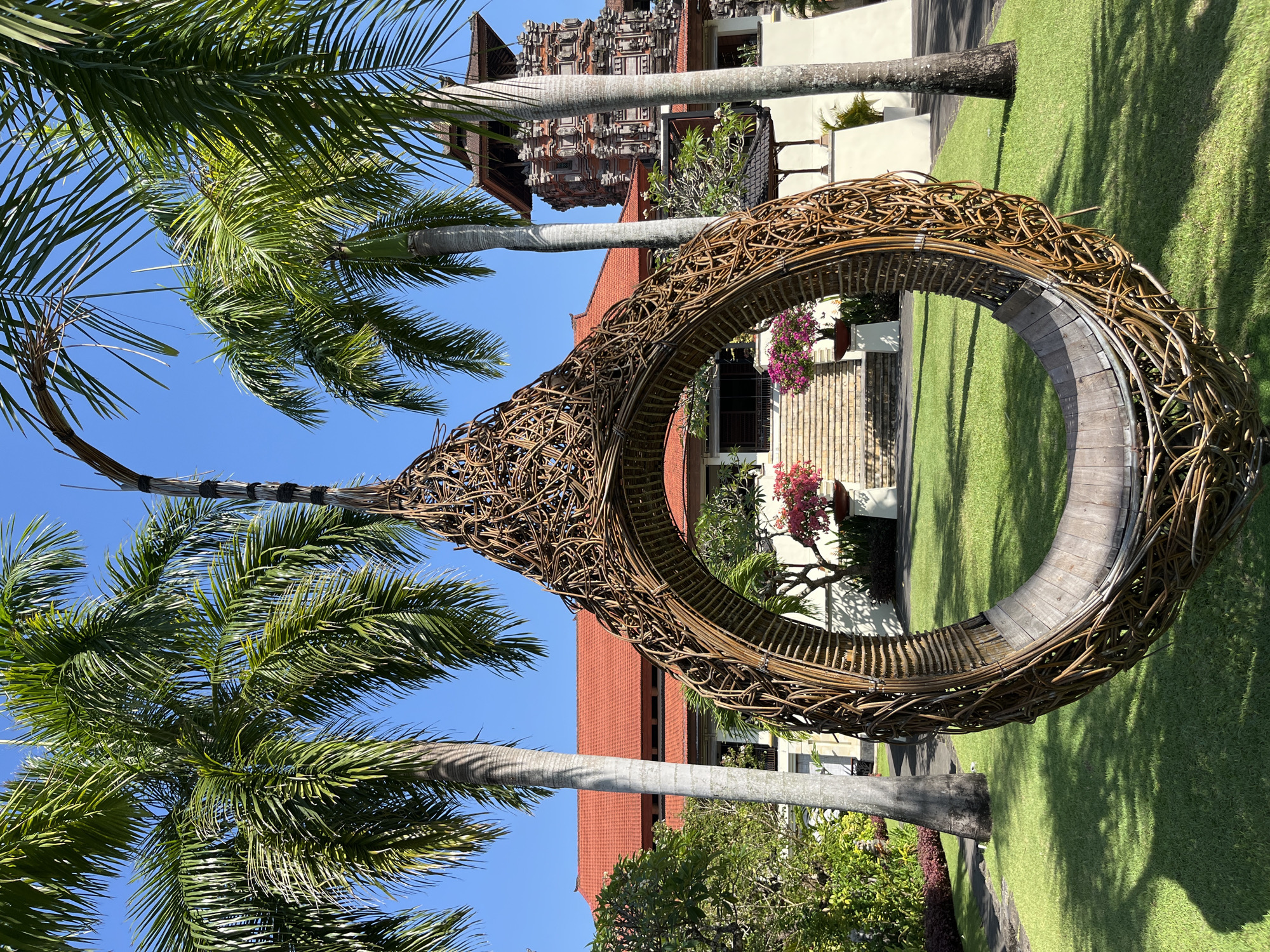 In Kuta Selatan, Indonesia, a woven egg-shaped swing with a wooden seat is positioned on a lush green lawn. Tall palm trees frame the scene, with their fronds reaching towards a clear blue sky. Behind the swing, a building with a red-tiled roof is visible, and to the right, a traditional Balinese tower with intricate stonework stands prominently. The architecture suggests a resort or cultural complex. The sun casts shadows on the grass, indicating a bright, daytime setting. There are no people or animals present in the image.
