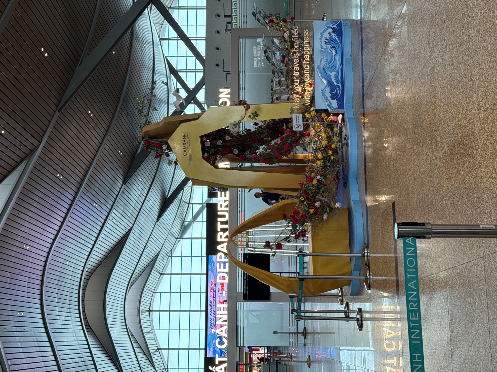 The image shows the interior of an airport terminal, specifically the departure area, with a decorative display in the foreground. The display features a golden arch with a tree-like structure covered in red and yellow roses. Below the arch is a boat-shaped sculpture adorned with fish figures and lights, resting on a blue base depicting waves. A sign next to the display reads, "May your travels be filled with joy and happiness."
In the background, large digital screens display text in Vietnamese and English. One screen prominently shows "ĐIỂM CẢNH DEPARTURE". Other signs and airport infrastructure like check-in counters and stanchions with velvet ropes are visible. The ceiling is characterized by a modern, curved architectural design with metal beams and lighting. A man in a dark shirt can be seen standing near a check-in counter in the middle ground. The floor is polished, reflecting the overhead lights and the decorative display. The overall environment suggests a busy international airport, likely Cam Ranh International Airport in Bắc Cam Ranh, Vietnam, given the presence of "CAMRANH INTERNATIONAL TERMINAL" on the display.