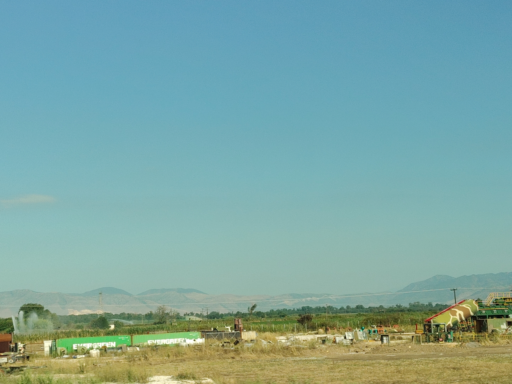 This image depicts a rural landscape under a clear blue sky. In the foreground, dry, yellowish-brown grass covers the ground. Behind this, a line of green shipping containers is visible, some bearing the word "EVERGREEN" in white lettering. Several industrial structures, including what appears to be a large water tower or tank with camouflage-like patterns, are scattered among the containers and surrounding vegetation. In the middle ground, a field of green crops, possibly corn, stretches towards distant hills. A water sprinkler is spraying water into the air in the left-center of the image, creating a misty effect. The background features a range of hazy, layered mountains. The scene appears to be located in a semi-industrial or agricultural area, likely on the outskirts of a city. There are no people visible in the image.