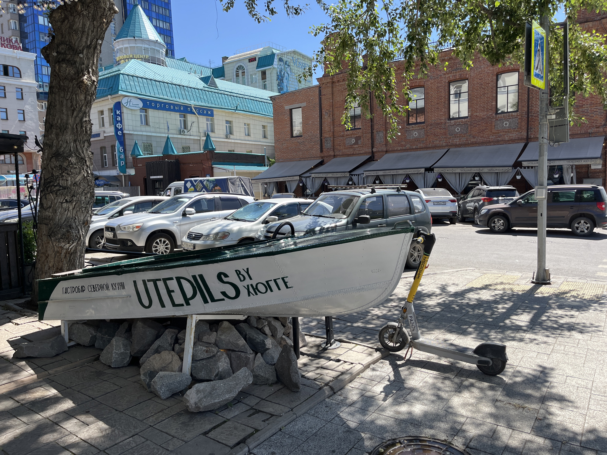 This media file is a photograph of a street scene in a city. The main subjects are a white boat displayed on a stone base, a yellow electric scooter parked next to it, and several cars parked on the street. In the background, there are buildings with a mix of architectural styles, including modern high-rise buildings and older brick structures with awnings.
The boat has text written on its side in dark green letters. On the left side, it reads "ГАСТРОБАР СЕВЕРНОЙ КУХНИ" (Gastrobara of Northern Cuisine), and to the right, it says "UTEPILS BY ХЮГГЕ" (Utepils by Hygge). The scene appears to be set during the daytime, with bright sunlight casting shadows from trees and buildings onto the paved ground. The weather seems clear and sunny.
Visible text includes the boat's inscription, as well as signage on the buildings in the background, such as "ДЕЛОВОЙ ЦЕНТР" (Business Center), "торговый пассаж" (shopping arcade), and "кафе бар" (cafe bar). There is also text on the electric scooter ("PA2474") and on license plates of the cars. The overall impression is one of an urban environment on a sunny day, with the boat and scooter adding a touch of novelty or artistic installation to the commercial streetscape. There are no people clearly visible in the image.