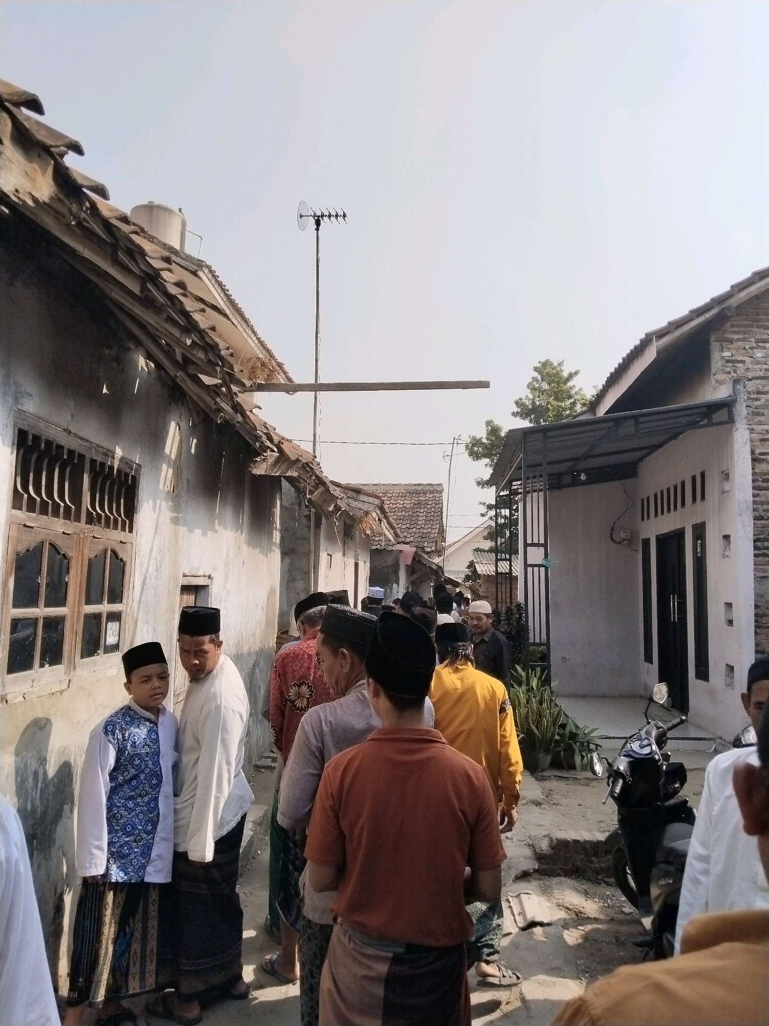 The image depicts a street scene in the city of Serang, Indonesia, with several individuals lined up. The architecture suggests a rural or semi-urban environment, characterized by older buildings with tiled roofs and white-washed walls. Some of the structures appear worn and in disrepair, with visible damage to the roofing. A water tank is visible on one of the rooftops. The individuals in the foreground and mid-ground are mostly men, many of whom are wearing traditional Islamic attire, including peci (caps) and sarongs. One young boy in the foreground is wearing a white shirt with a blue patterned vest and a sarong. The group appears to be walking down the street, suggesting a procession or a gathering. In the background, there is a motorcycle parked on the side of the street, and a television antenna is mounted on a pole. The sky is bright and clear, indicating daytime. The overall atmosphere is that of a community event or daily life unfolding in a local Indonesian setting.