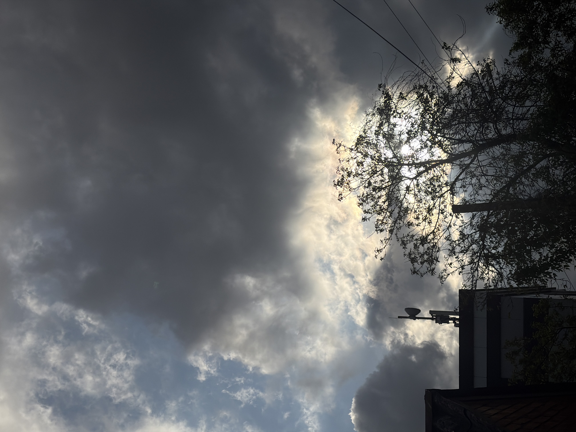 The image depicts a cloudy sky above a building with trees silhouetted against the light. The sky is filled with dark, heavy clouds on the left and lighter clouds with patches of blue sky on the right. In the upper right, tree branches partially obscure a bright light source, likely the sun. A building is visible in the lower right, with a weather instrument mounted on its side. Powerlines run along the top of the image. The location is unknown and unconfirmed.
