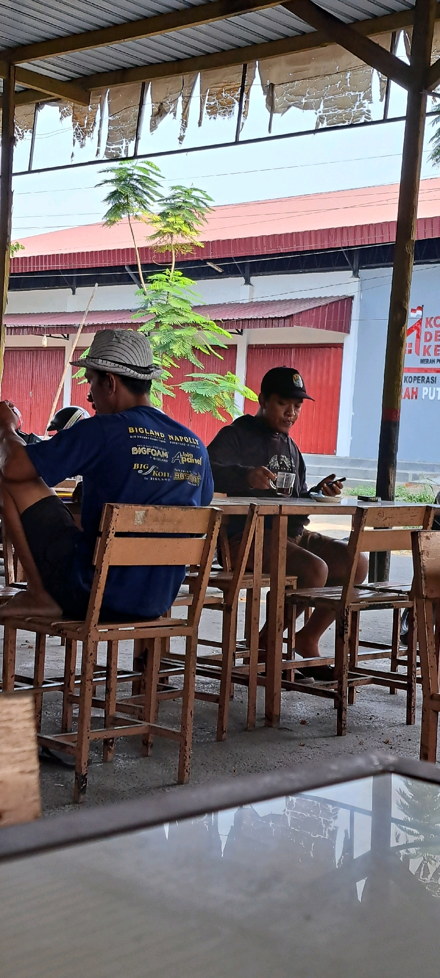 Two men are seated at a table outdoors, likely at a cafe or warung in Nogosari, Indonesia. The man on the left wears a blue t-shirt with text and graphics, a striped hat, and black shorts, and is seen with his left arm on the table. The man on the right wears a black hoodie, a black baseball cap, and is holding a phone and a glass of dark liquid, appearing to be drinking. They are seated on wooden chairs at a wooden table. The background shows a building with a red facade and a white section with text. The overhead structure is made of corrugated metal and wooden beams, with some of the ceiling material torn and hanging. Green foliage is visible in the background. The ground is a rough, concrete surface.