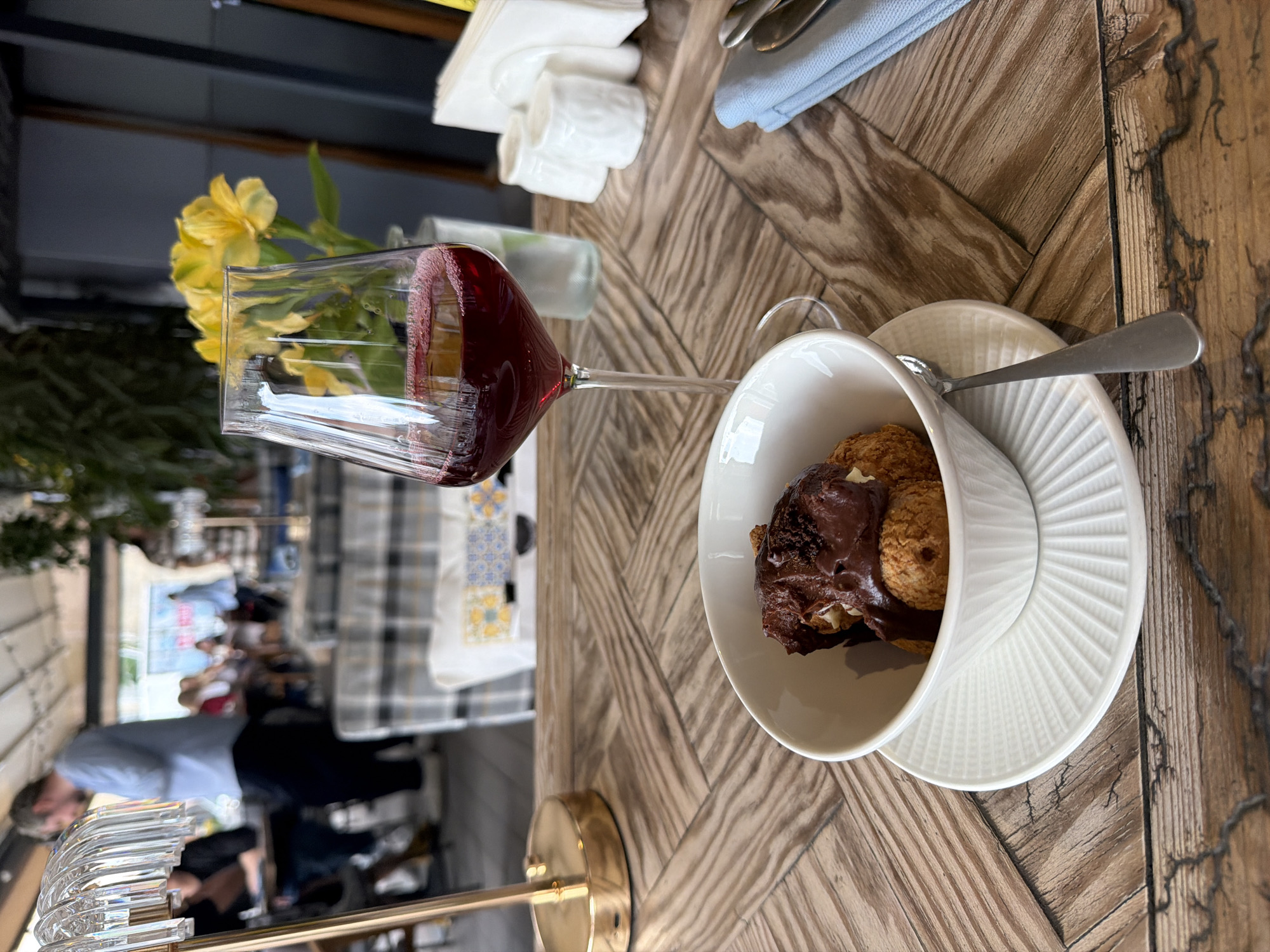 A table in what appears to be a restaurant is the main focus of the image. The table has a herringbone-patterned wooden surface. A glass of red wine is positioned in the top left corner of the image, with yellow flowers behind it in a glass. To the right of the wine glass, there's a stack of neatly folded white napkins and a blue napkin.
A white bowl filled with what looks like pastries covered in chocolate is placed on a white saucer. A silver spoon rests on the saucer, near the bowl.
In the background, an open space with tables and chairs can be seen. People are seated at some of the tables. Through the open area, there are glimpses of what might be an outdoor setting. The scene is presumably in Moscow, Russia.