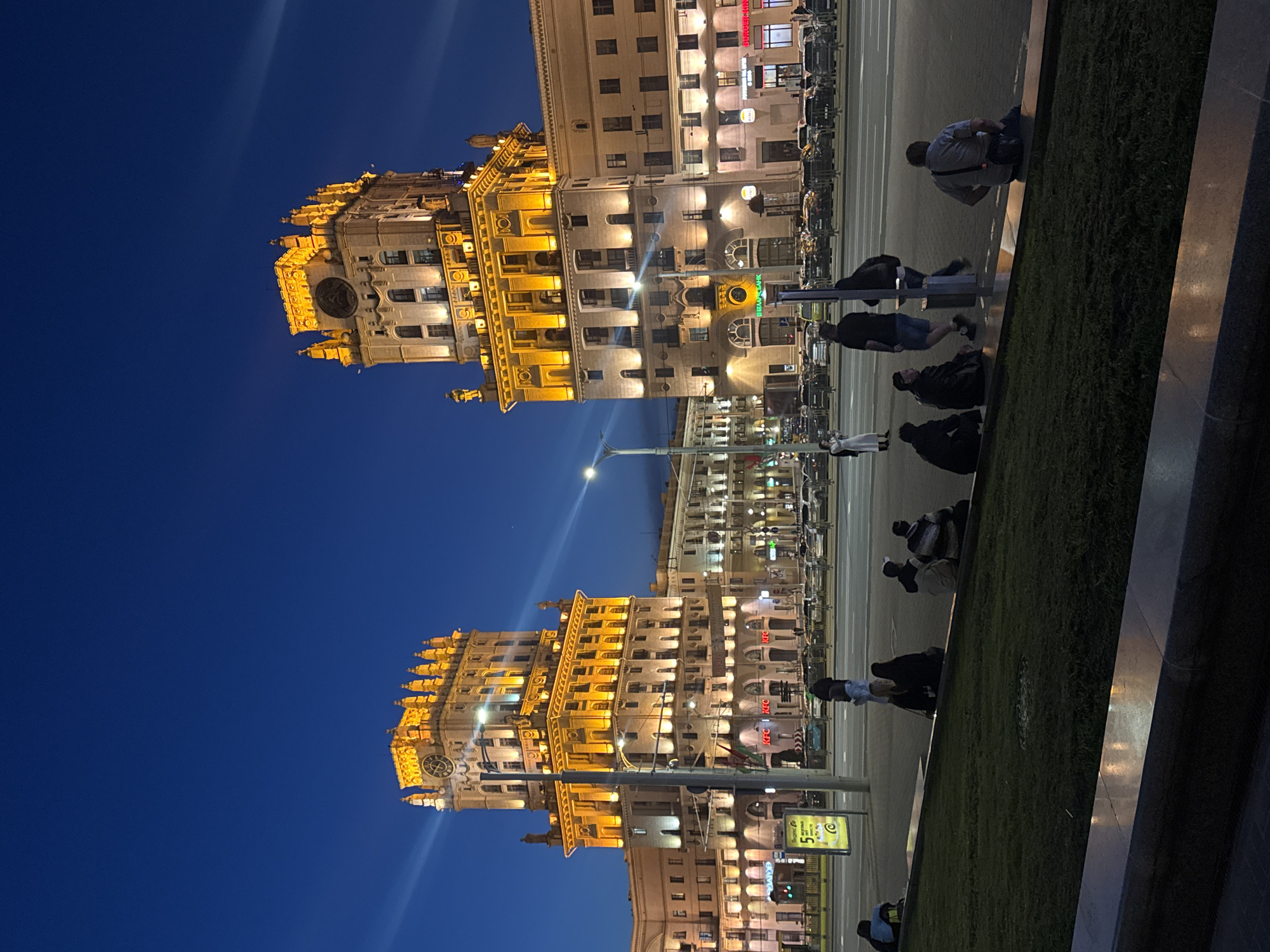 The image shows the Independence Avenue in Minsk, Belarus, at night. The iconic twin buildings, illuminated in warm light, are visible against a dark blue sky. The street scene includes a sidewalk with people walking and sitting near a green grassy area. Cars are parked along the street. Streetlights and signs are present, adding to the urban environment. The photograph is taken from a low angle, emphasizing the height of the buildings.