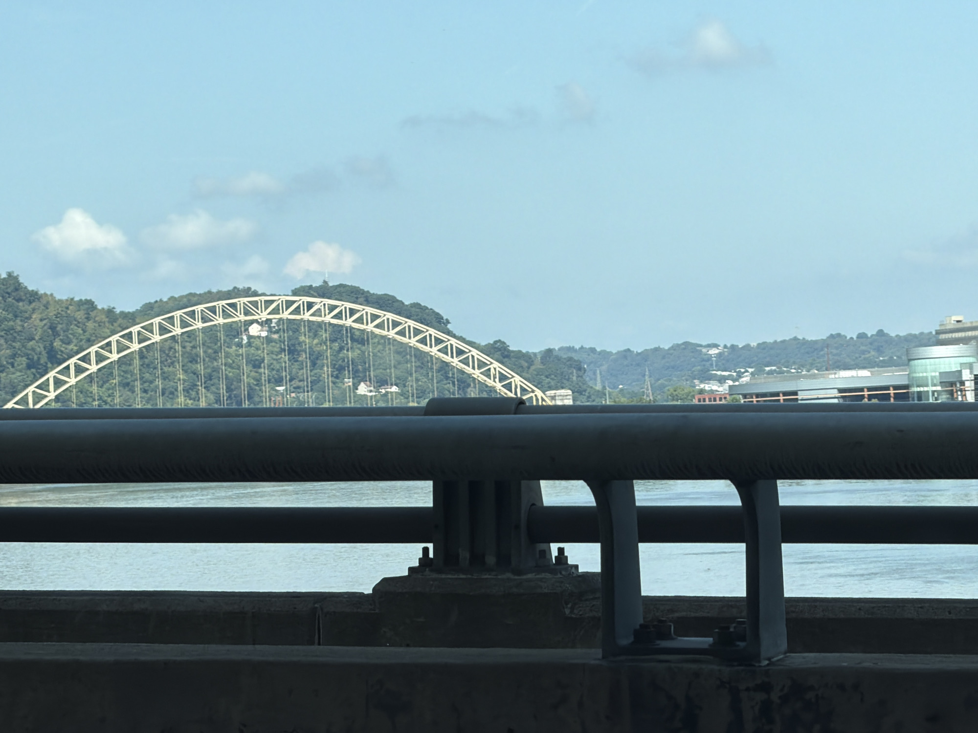 This is a photograph taken from a bridge overlooking the Ohio River in Pittsburgh, Pennsylvania. The foreground of the image is dominated by the dark, industrial structure of the bridge itself, with thick metal railings and supports obscuring the view. Beyond the bridge, the Ohio River stretches out, its surface calm and reflecting the light blue sky. In the middle ground, a large, arched bridge, likely the Fort Pitt Bridge, is visible spanning the river. The bridge is a light color, possibly yellow or white, and its intricate latticework is clearly defined against the green, tree-covered hills that form the background. Several buildings, including a cylindrical glass structure, are visible on the far bank of the river, nestled among the trees. The sky is a clear, bright blue with a few scattered white clouds. The overall impression is one of a typical urban river landscape on a sunny day.