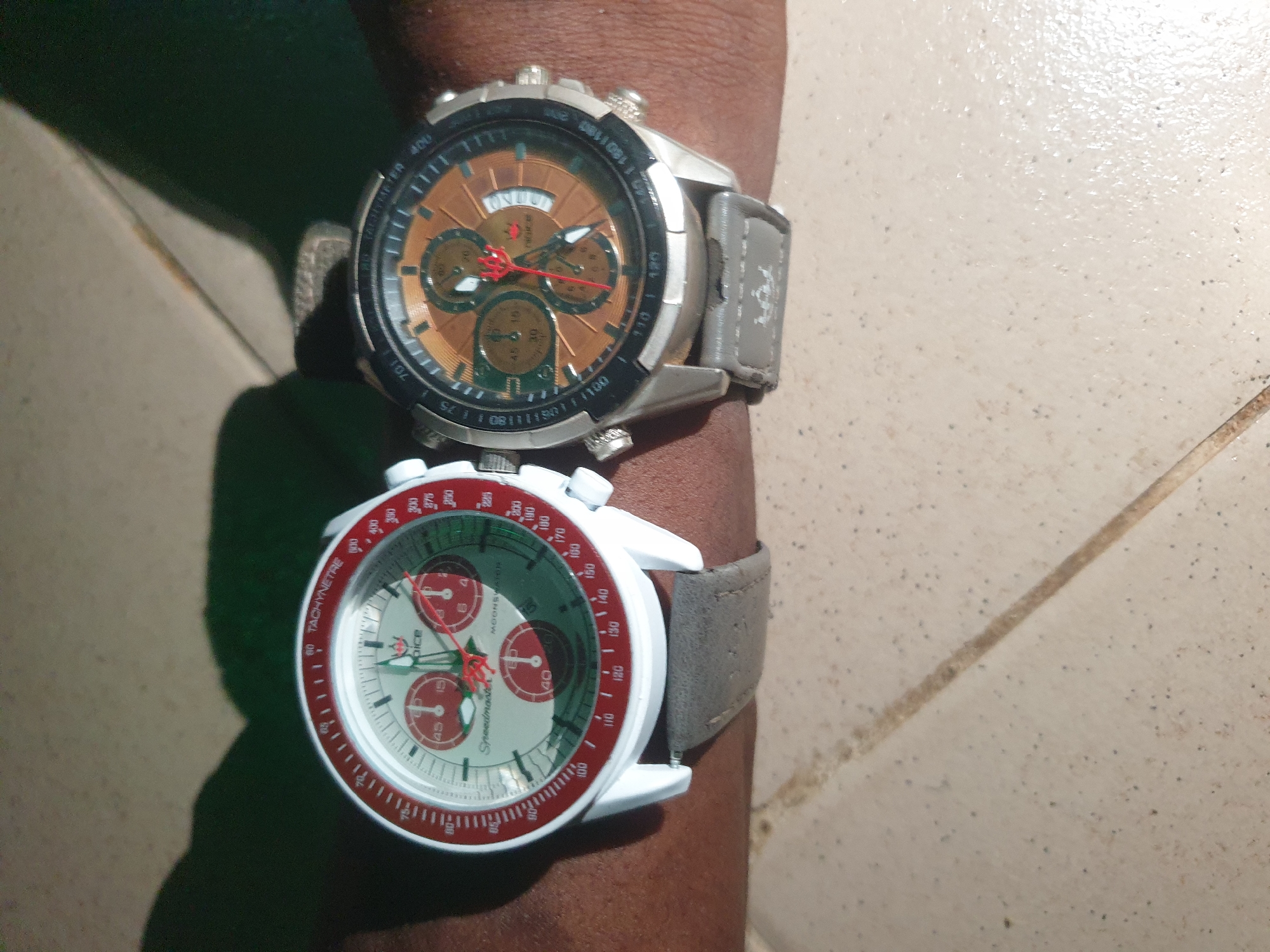 Two wristwatches are displayed side-by-side on a person's arm. The watch on the left has a white face with red accents, featuring a tachymeter bezel and three sub-dials. The watch on the right has a gold-toned face with red accents, also with a tachymeter bezel and three sub-dials. Both watches appear to have grey leather straps. The background is a mottled surface, possibly a tiled floor, with shadows cast from the watches and arm. The image quality is moderate, with some blurring and noise. The location is identified as Anilelerin, Nigeria.
