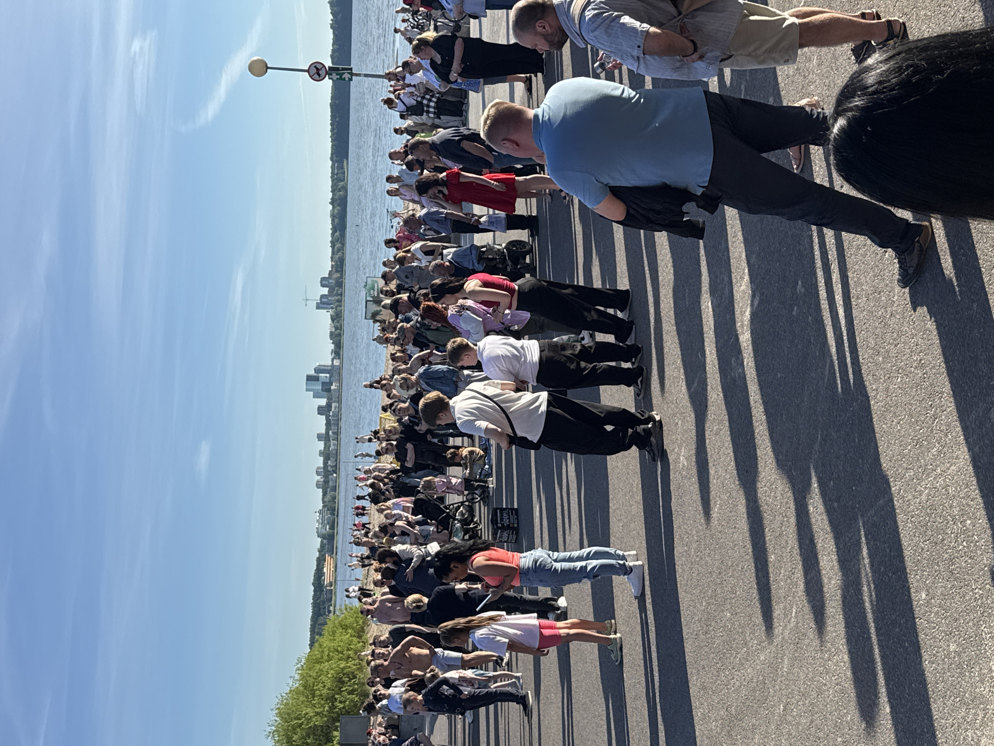 An outdoor gathering of approximately 100 people, predominantly adults, is taking place on a paved area adjacent to a body of water, likely the Baltic Sea, with a city skyline visible in the distance. The scene is illuminated by bright sunlight, casting long shadows across the ground. The crowd is diverse in age and attire, with many dressed in casual summer clothing. Several individuals are standing, some in small groups, while others are walking or observing something out of frame. A few children are present, and one person appears to be in a wheelchair. In the background, a sandy beach area is visible with more people, and a line of trees separates the beach from the water. The city skyline features modern high-rise buildings and construction cranes. A lamppost with a circular sign featuring a dog symbol and a circle with a line through it is positioned on the right side of the frame, indicating a no-dogs-allowed zone. The overall impression is of a public event or casual social gathering on a pleasant day in Tallinn, Estonia.
