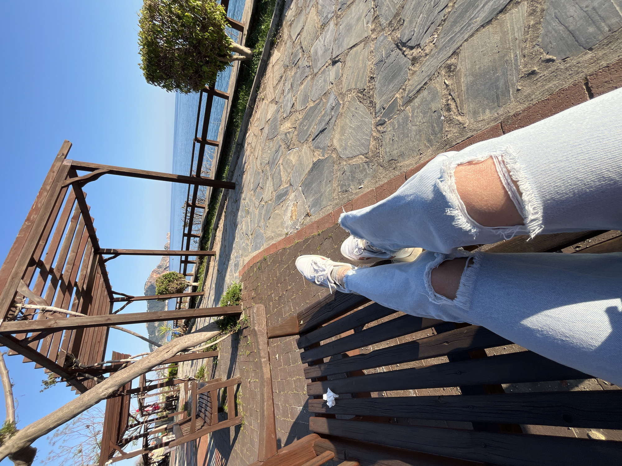 The image captures a first-person perspective of someone relaxing on a dark brown wooden bench at a scenic coastal location in Alanya, Turkey. The primary subject is the person's legs, which are stretched out on the bench, wearing light blue distressed jeans with rips at the knees and white athletic shoes with white laces.
The bench is part of an outdoor promenade or overlook. The ground immediately surrounding the bench is paved with irregular gray stone slabs, bordered by a reddish-brown brick line. Adjacent to the stone paving, a narrow strip of green grass and low shrubs runs parallel to a wooden fence or railing. Beyond this fence, the calm, deep blue sea stretches out to meet a clear, bright blue sky on the horizon. A distant, rugged landmass or mountain is visible across the water.
To the left, a substantial wooden pergola or trellis structure provides architectural interest, featuring robust wooden beams and supports, some of which appear to be naturally growing tree branches integrated into its design. A small, round, green tree is also visible near the wooden fence by the sea. Bright sunshine illuminates the scene, casting sharp, distinct shadows across the bench and the ground, suggesting it is daytime, likely late morning or early afternoon on a clear, sunny day. A crumpled white tissue is visible on the wooden bench beside the person's right leg. The overall mood is tranquil and serene, inviting relaxation and enjoyment of the beautiful coastal view. There is no visible text in the image.