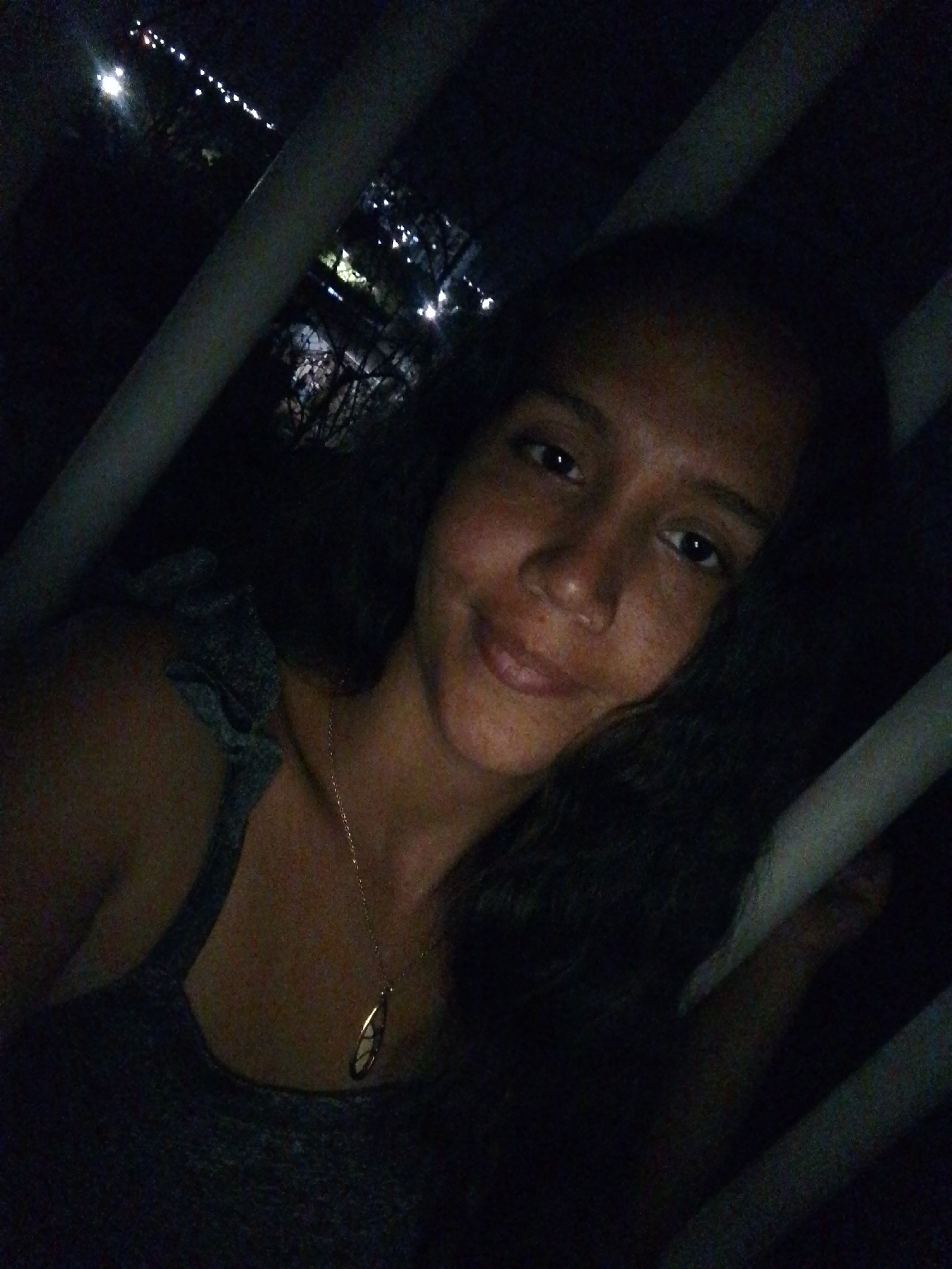 The image depicts a young woman posing for a photograph at night in Juan Griego, Venezuela. The woman, with dark hair and a light complexion featuring visible freckles, occupies the central and lower-right portion of the frame, looking directly towards the viewer with a slight smile. She is wearing a dark, possibly gray, top with a textured or ruffled detail on her right shoulder strap. A delicate gold-colored chain necklace with an oval pendant is visible around her neck. Multiple light-colored, elongated elements, appearing as bars or rails, traverse the foreground diagonally and vertically, partially obscuring the woman and the background. Through the gaps in these foreground elements, a distant urban landscape is visible in the background, characterized by numerous small, bright city lights against a dark sky, confirming the nighttime setting. The primary illumination source is directed towards the woman's face, casting the majority of the scene in shadow.