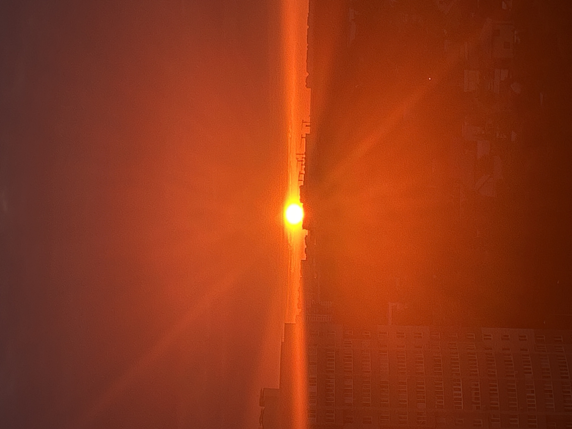 The image captures a sunset over the city of Ivanteyevka, Russia. The sky is dominated by vibrant hues of orange and red, with the sun a bright, luminous orb near the horizon. Rays of sunlight emanate from the sun, creating a dramatic effect across the sky. In the foreground, a tall, multi-story building with numerous windows is visible on the left side of the frame. Silhouettes of other buildings and construction cranes can be seen in the distance, indicating an urban environment. The overall atmosphere is serene and visually striking due to the intense colors of the sunset. There are no people or animals visible in the image.