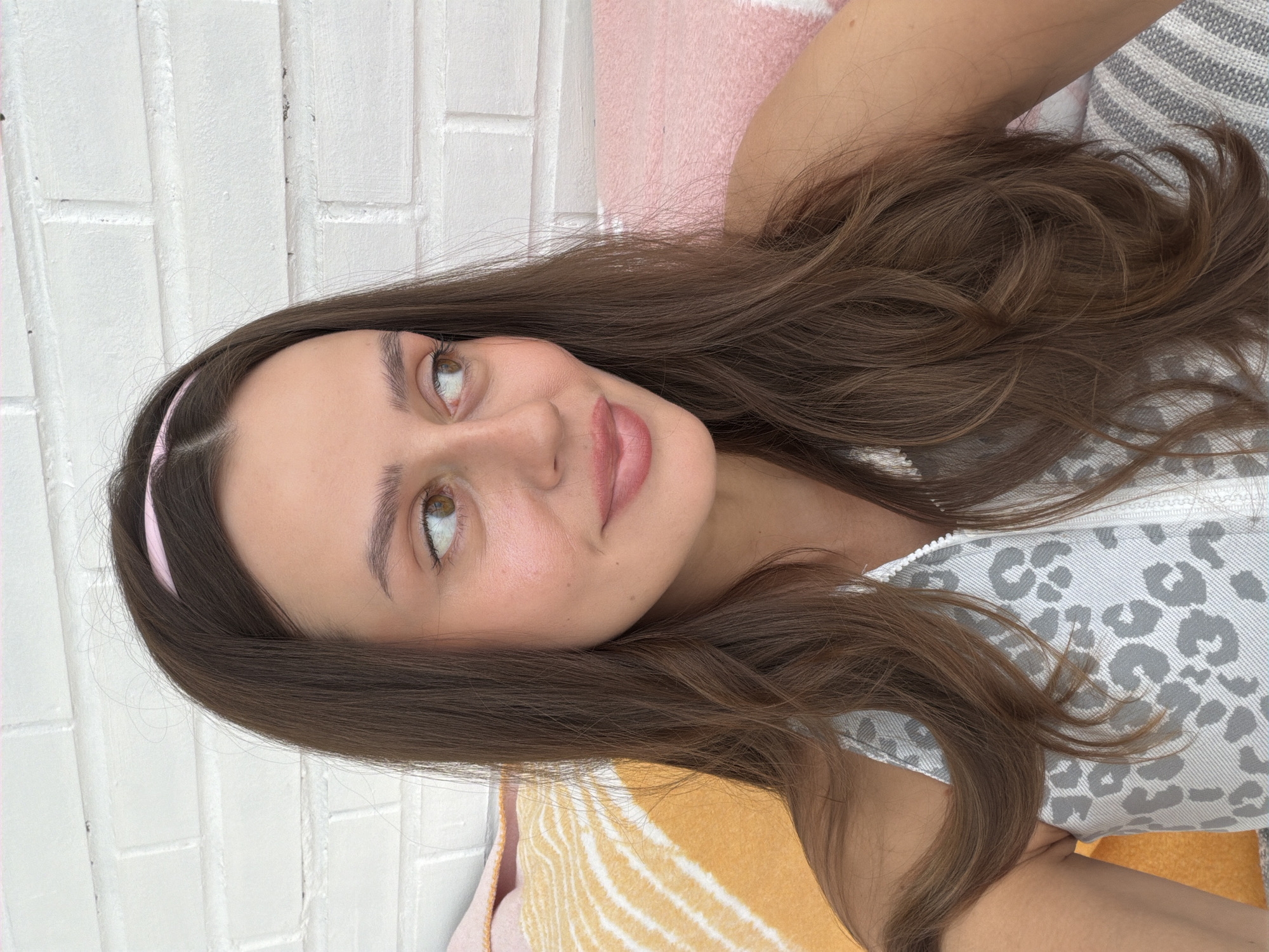 The image shows a close-up, waist-up shot of a woman with long, brown hair, wearing a white sleeveless top with a gray leopard print. She has a pink headband on and is looking to her right, with her lips slightly parted. She is seated on a bed with pillows in the background, one orange with yellow stripes and another pink. Behind the bed, a white brick wall is visible. The scene appears to be indoors, likely in a residential setting, possibly in Krasnoyarsk, Russia, given the provided context, though there are no specific visual cues to confirm this location. The lighting is soft and natural, suggesting it was taken during the day.