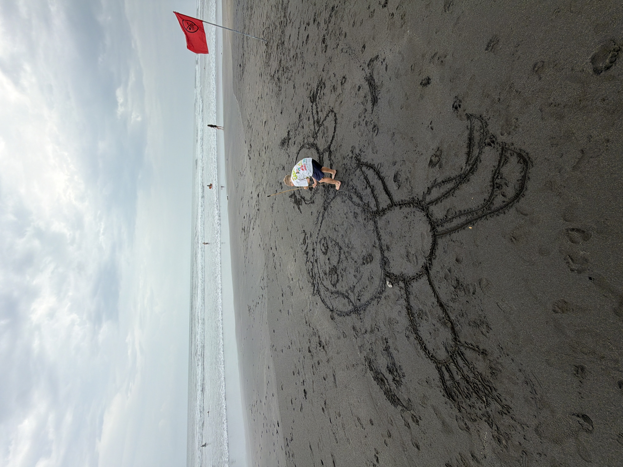 A low-angle, full shot captures a child drawing a large smiley face in the dark sand on a beach in North Kuta, Indonesia. The child, wearing a white t-shirt with colorful graphics and dark blue shorts, crouches down and uses a stick to draw the design. The sandy beach extends towards the ocean, where gentle waves crash. In the distance, a red flag is planted in the sand, and a few people are visible in the water. The sky is overcast with dramatic clouds.