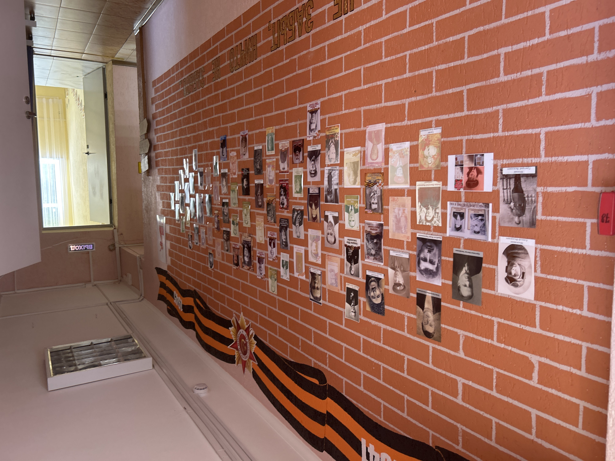 The image shows a hallway with a brick wall decorated with portraits of people and a red and orange ribbon with a star. The wall has the inscription "НИКТО НЕ ЗАБЫТ, НИЧТО НЕ ЗАБЫТО" which translates to "No one is forgotten, nothing is forgotten". There are also several doors, one of which is open, revealing a room with a window and curtains. The overall scene suggests a memorial or a place of remembrance. The inscription and the decorations are typical of memorials related to World War II in Russia, celebrating Victory Day. Gelendzhik, a resort city in Krasnodar Krai, Russia, is known for its numerous monuments and memorials dedicated to the Great Patriotic War.