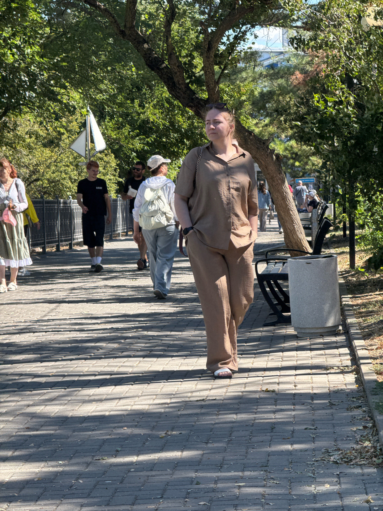 A group of people are walking on a paved pathway in a park-like setting. In the foreground, a woman in a tan linen shirt and pants walks away from the camera, with her left hand in her pocket. To her left, other individuals are walking in the same direction. One woman in a white dress with a pink scarf, a man in a black t-shirt and shorts, and a person in a white hoodie and light blue jeans are visible. Further down the path, more people are walking, some sitting on a bench, and a trash can stands near the pathway. The path is bordered by a railing and trees casting shadows on the ground, which is covered in fallen leaves. In the background, a white sail-like structure is visible among the trees. The scene appears to be in Anapa, Russia.