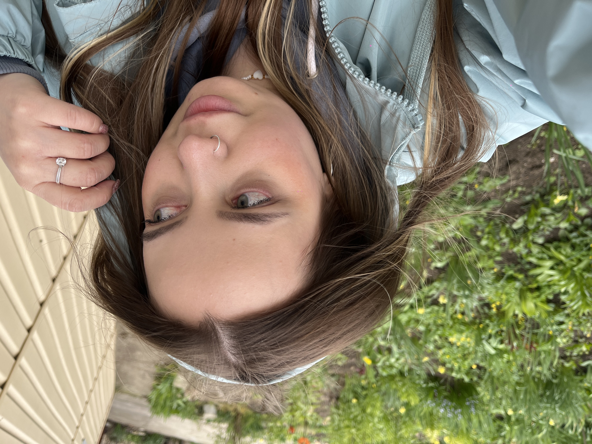 A young woman with brown hair, fair skin, and a nose ring is holding her hand up showing a ring on her finger, while posing for a selfie. She is wearing a light blue jacket and a necklace. Behind her, there is a green grassy area with small yellow flowers. To the left, a light-colored wooden structure is visible. The image does not provide enough information to determine its location in Novopokrovskaya, Russia.