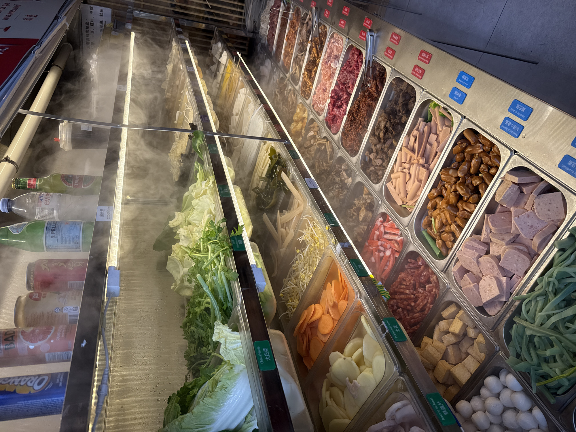 The image shows a food display case, likely in a self-service restaurant or market. The case contains a variety of ingredients commonly used in hot pot or similar Asian cuisine. The left side of the display holds fresh produce, including leafy greens (Chinese cabbage), cilantro, bean sprouts, sliced carrots, and other vegetables. A light strip runs along the top, and there is mist or condensation visible.
The right side of the display features various prepared ingredients, such as surimi (crab sticks), sausages, processed meats (cubed ham), sliced potatoes, tofu cubes, lotus root slices, and other items. Small labels with Chinese and possibly French text identify the ingredients. Bottles of San Pellegrino water, Tsingtao beer, and other beverages are seen in the background. The overall setting suggests an Asian restaurant or food market. The OCR text hints it is likely in Paris, France.