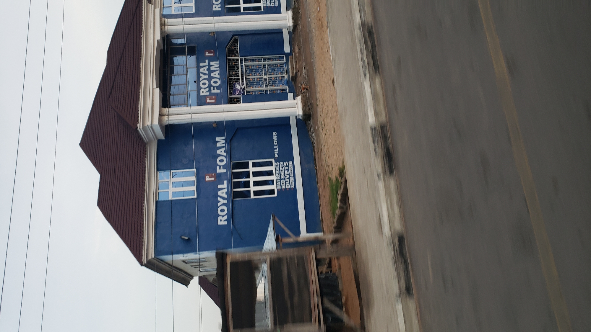 The image shows a blue building with the text "ROYAL FOAM" in white letters. The building has a dark brown roof and white trim around the windows and columns. There are several windows on the building, some of which are large and rectangular. Below one of the windows, there is a list of products: "MATTRESSES BED SHEETS DUVETS". To the left of the building, there is a wooden structure with several shelves, some of which are covered with netting. The building appears to be located on a street in Jalingo, Nigeria, with a road in the foreground. The sky is overcast.