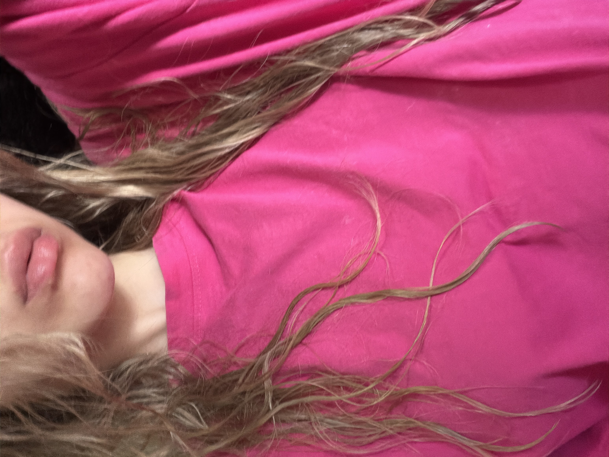 A close-up image shows a person lying down, wearing a bright pink shirt. The person's face is partially visible on the left side of the image, featuring lips and a portion of the neck. Long, light brown hair strands are scattered across the pink shirt. The background is dark and indistinct. The location and city are unknown.