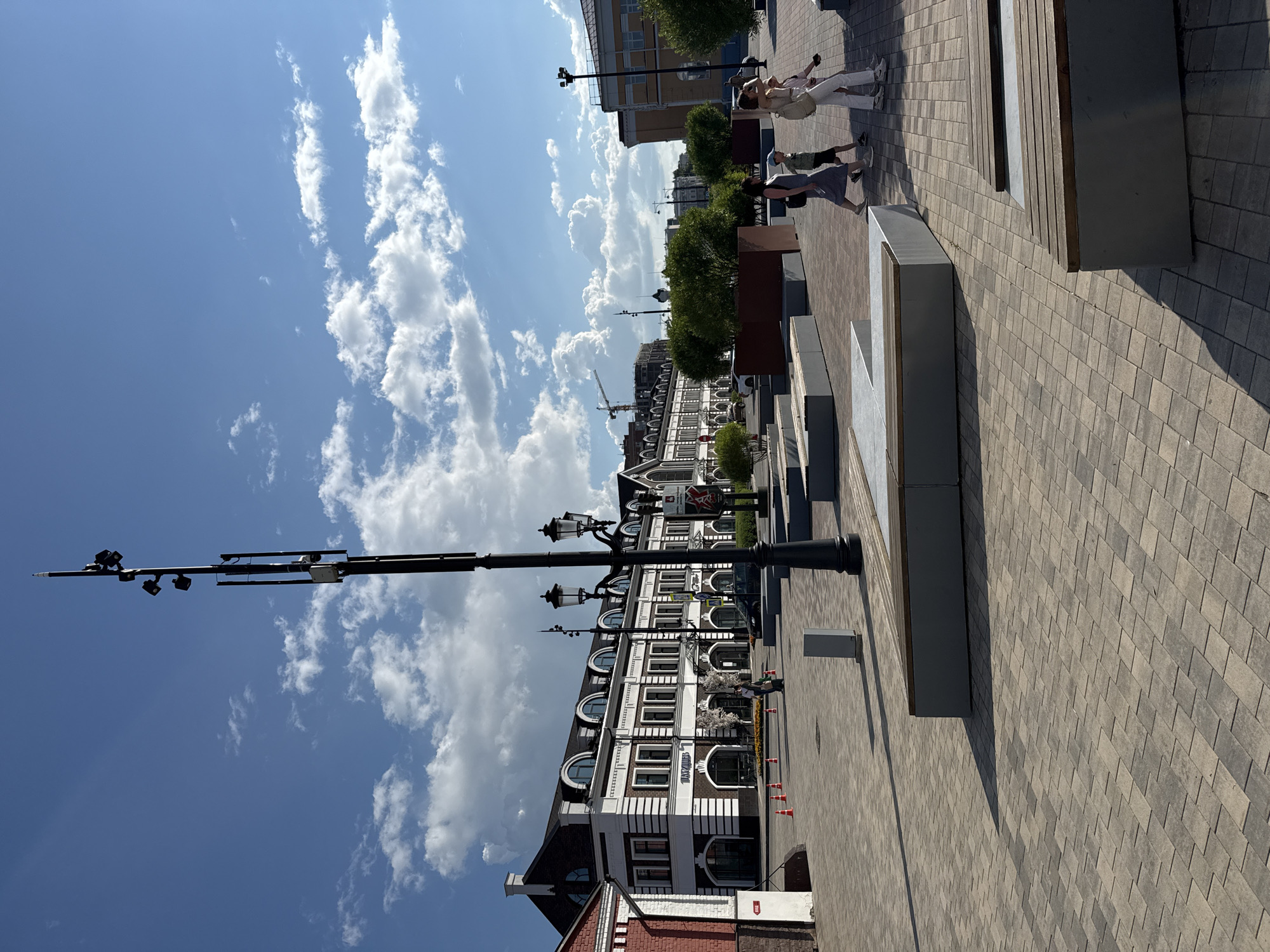 The image depicts a city square in Perm, Russia, featuring a modern architectural style with a blend of old and new elements. The foreground is dominated by a paved area with patterned stone tiles, interspersed with contemporary concrete benches and planters. A tall, black lamppost with multiple lights and what appears to be communication equipment stands prominently in the mid-ground.
In the background, a long, white and dark-trimmed building with arched windows and mansard roofs suggests a historical structure that has been renovated or is part of a development project. To the left, a red brick building adds a touch of older architecture. Several people are walking across the square. A family, consisting of two adults and two children, are walking from right to left. Another person is walking towards the right. Orange traffic cones are visible near the building on the left, indicating a possible temporary closure or construction work. Green trees in planters are strategically placed throughout the square. The sky is blue with scattered white clouds, and the sun casts distinct shadows on the paved ground. The overall scene conveys a sense of an urban public space designed for leisure and activity.