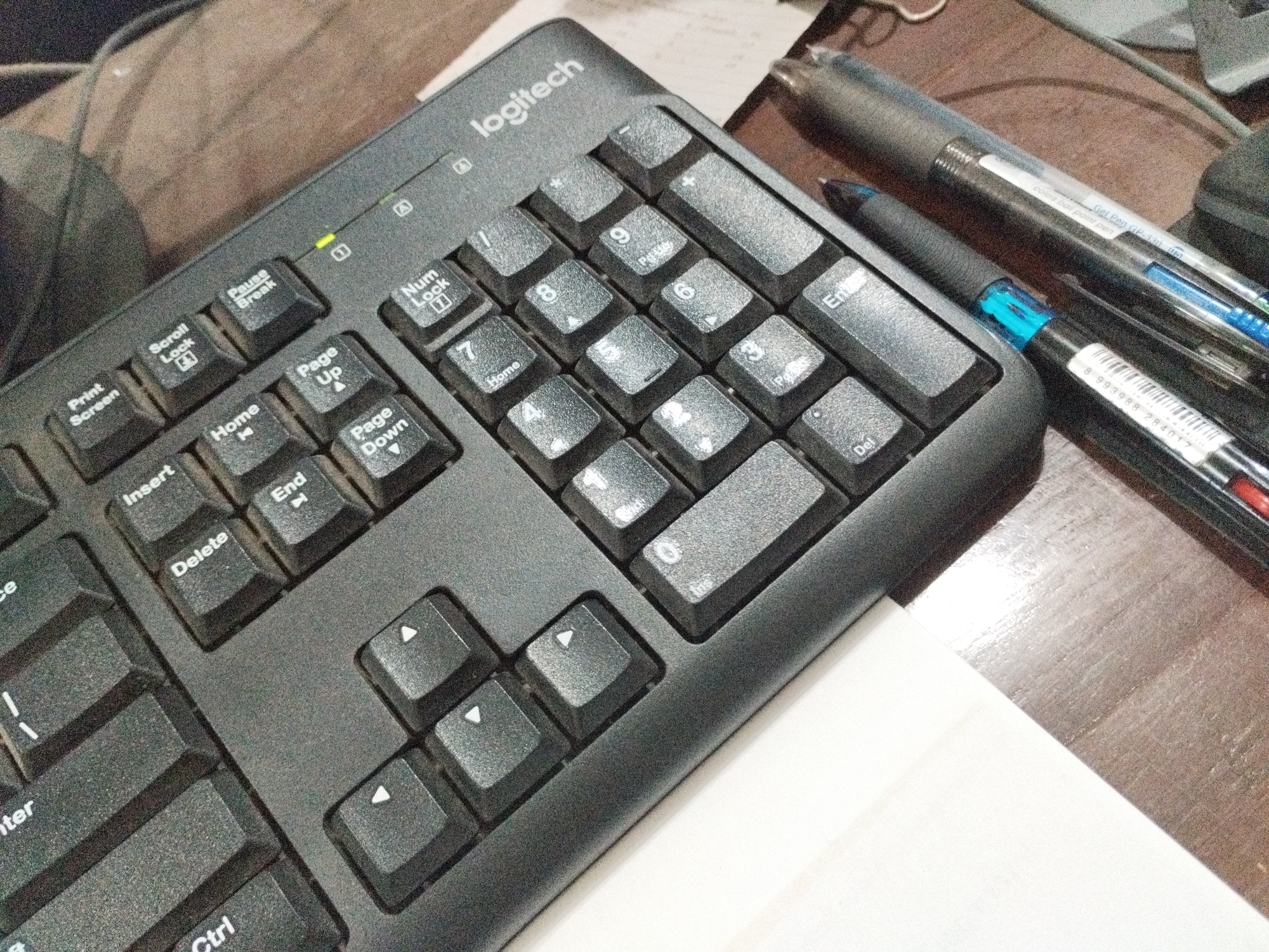 A black Logitech keyboard is positioned on a wooden surface, with a white piece of paper partially covering the surface. Two pens, one black and one with blue ink, are placed on top of the keyboard. The keyboard's keys are visible, including standard letter and number keys, as well as function keys like "Print Screen," "Scroll Lock," and "Pause Break." An indicator light, likely for Num Lock or Scroll Lock, is illuminated green on the keyboard. The scene suggests a workspace or desk setup. There are no people visible in the image. The location of Pehserut, Indonesia is not depicted or implied by any visual elements in the image.