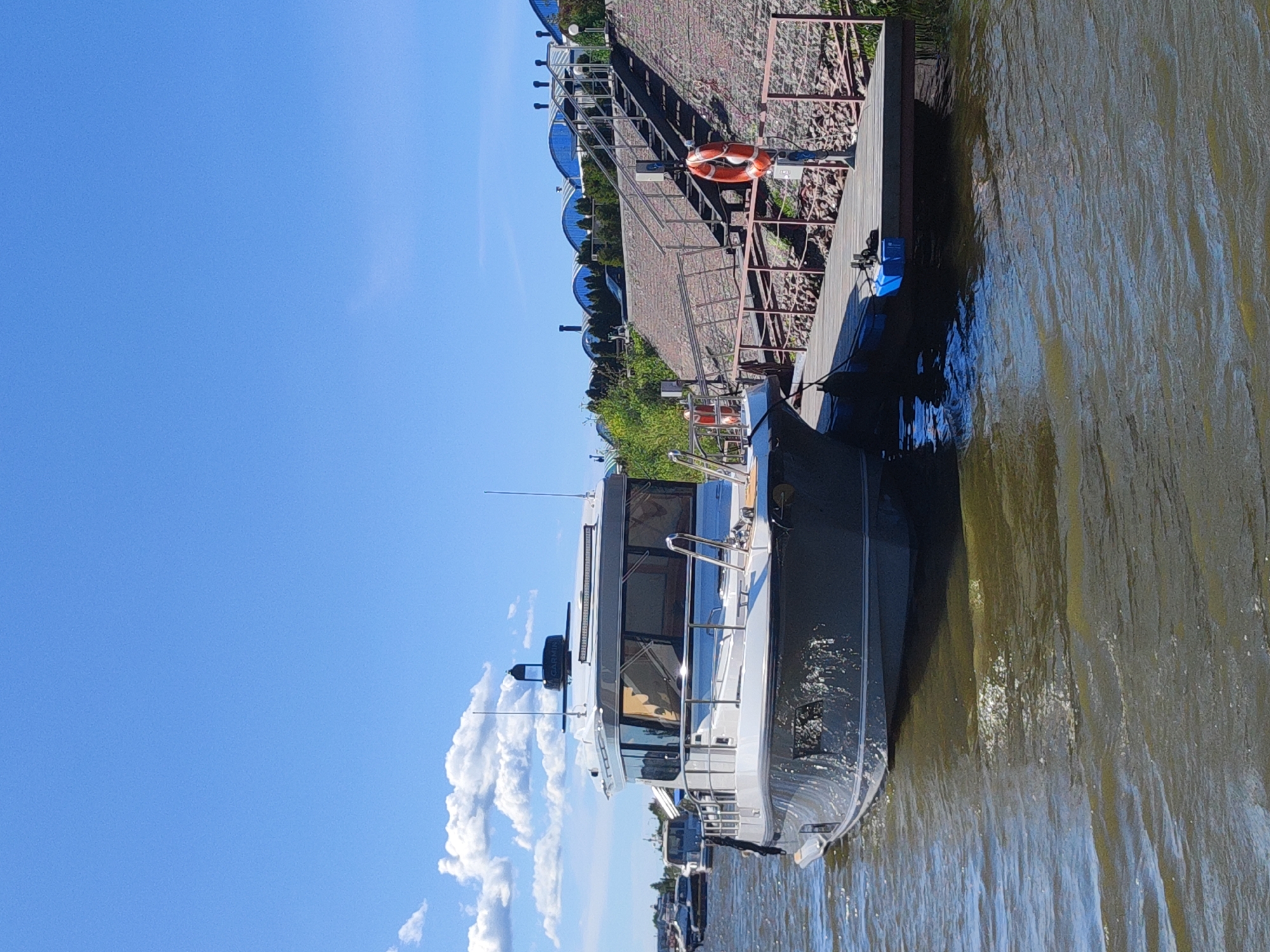 This is a vertical, eye-level shot of a modern, grey motor yacht docked beside a brick walkway and wooden pier. The yacht, with its sleek design and large windows, is the primary subject. It has a prominent radar dome on its roof, and a Garmin antenna is visible on the back. In the background, a lifebuoy is mounted on a railing. Beyond the pier, several domed structures with reflective blue roofs and lush green trees are visible, suggesting a developed waterfront area. The sky is a clear, vibrant blue with scattered white clouds. The water next to the yacht is a murky, greenish-brown, with ripples indicating slight movement. Other boats are moored further in the distance. The overall scene is bright and sunny, suggesting a pleasant day in a coastal or riverside urban environment, likely Saint Petersburg, Russia given the context. There are no people visible in the image.