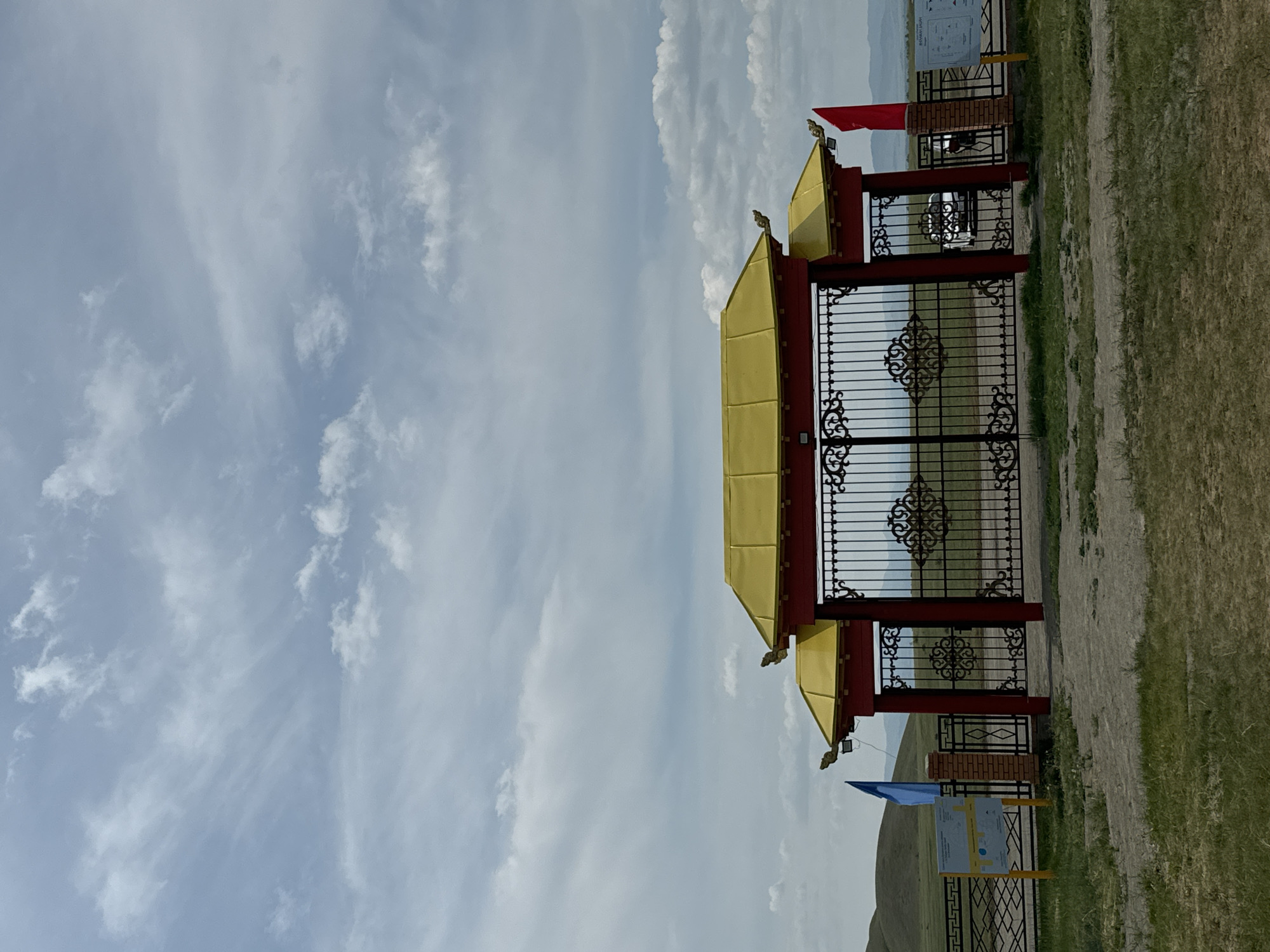 The image shows an ornate gate with red pillars and a golden, pagoda-style roof. The gate is made of black wrought iron with intricate scrollwork. On either side of the gate, there are brick pillars. A blue flag flies on the left side, and a red flag flies on the right side. Behind the gate, a white car is visible. The landscape behind the gate is open and hilly, with distant mountains under a cloudy sky. The foreground is a grassy area with a dirt path. Informational signs are posted on either side of the gate. The location is the entrance to the Orongoi Buddhist Temple complex in Orongoi, Russia.