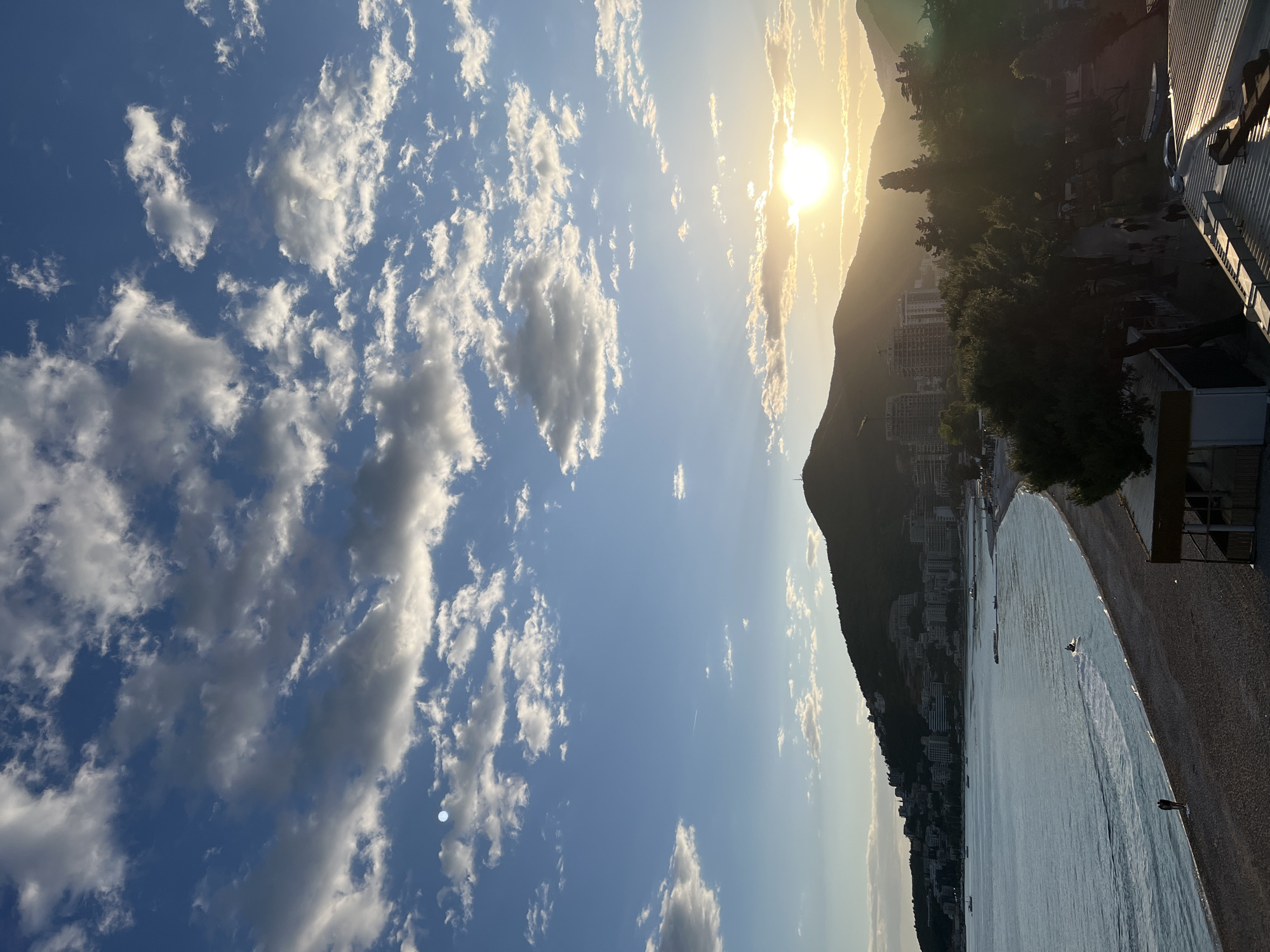 The image presents a scenic view of the city of Budva, Montenegro, during sunset. The sky is filled with white, fluffy clouds against a blue backdrop. The sun is positioned behind a mountain, casting a golden glow around it. A mountain range runs along the coastline, with residential buildings lining its base. The water is calm. The beach area is partially visible with a few people and boats. A building's architecture can be seen on the side.