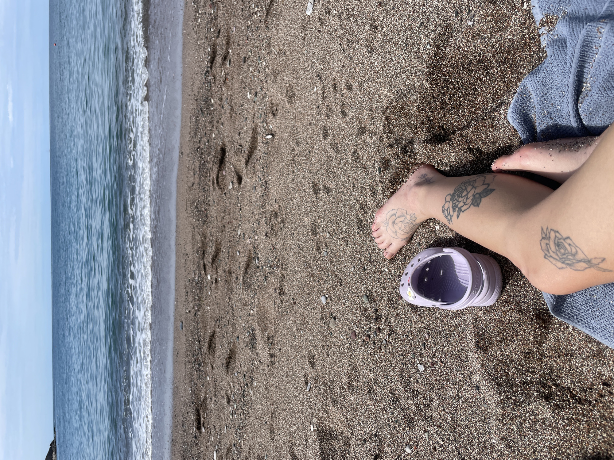The image depicts a beach scene in Rafailovići, Montenegro. The left portion of the frame shows the Adriatic Sea with gentle waves meeting the shoreline. The sand is dark and coarse, with visible footprints leading away from the water. To the right, a person is sitting on a blue towel, their legs extended. The person has tattoos of flowers, possibly roses, on their feet and legs. A pair of lavender-colored Crocs sandals is next to the person's leg. The overall scene is bright and appears to be taken during daylight hours.