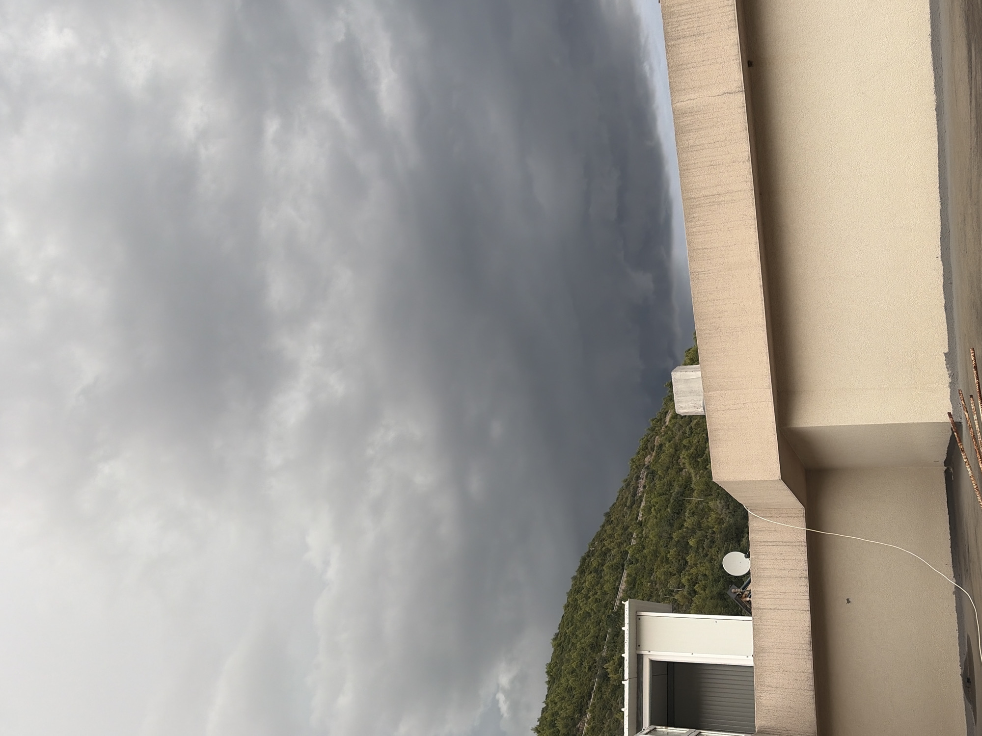 A cloudy sky looms over a building and a vegetated hillside in Budva, Montenegro. The sky is filled with dark, heavy clouds, suggesting an approaching storm. In the foreground, a multi-story building with beige walls and a light-colored parapet is visible. A white structure with an open door is situated on the roof of the building. A satellite dish is also present on the roof. A white cable snakes across the rooftop. The hillside to the left is covered with dense green vegetation.