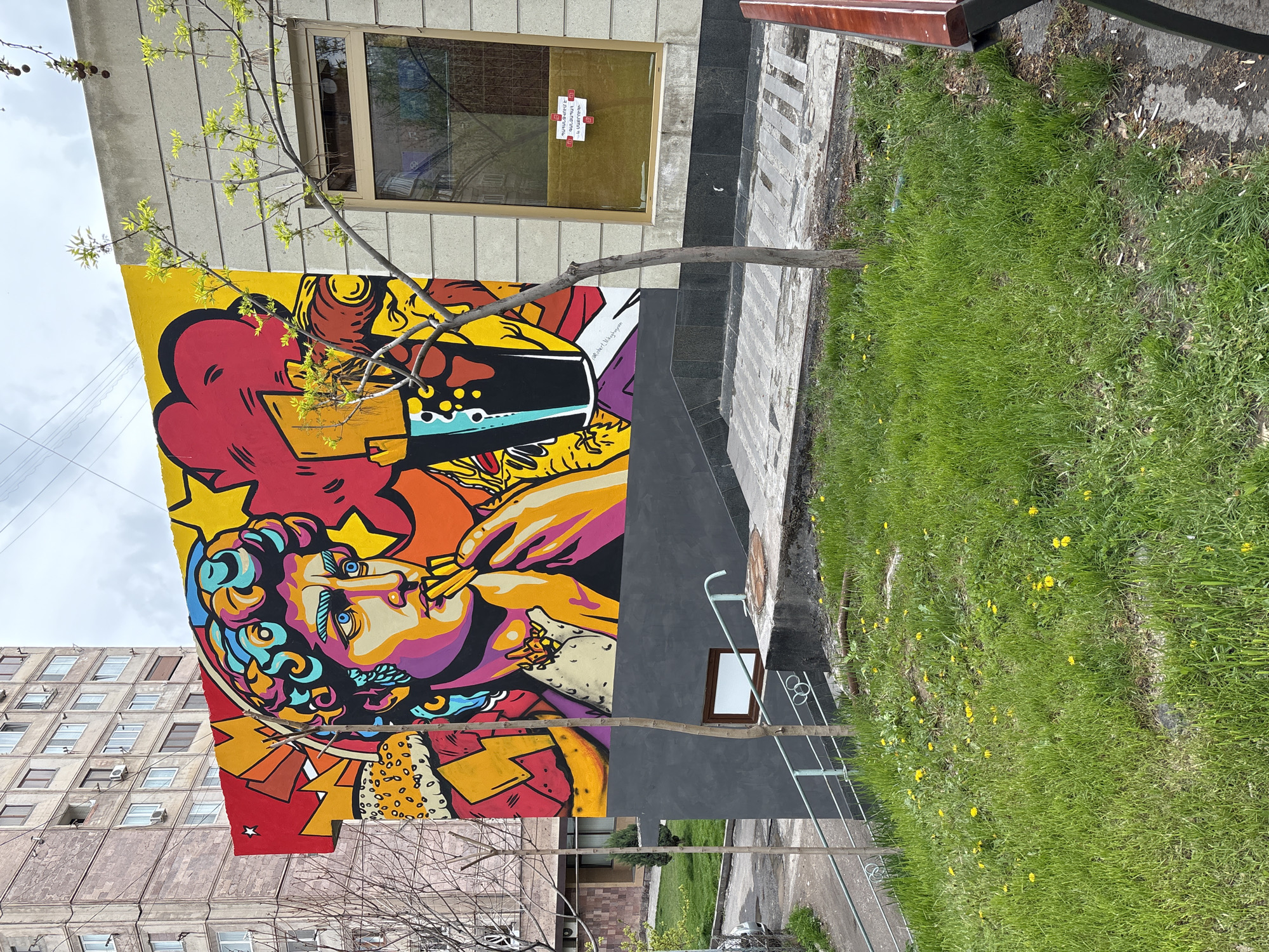 This vertical shot showcases a vibrant, multi-colored mural on the side of a building in an urban setting, likely in Yerevan, Armenia.
The central subject of the mural is a stylized, pop art-inspired depiction of a person's face, possibly a classical or mythological figure. This individual has striking blue eyes, a prominent nose, and elaborate, curly hair rendered in bright shades of blue, purple, orange, and yellow. They are shown with an object, possibly a pipe or a large straw, at their lips, from which a substantial red and yellow cloud of smoke or vapor emanates. The background of the mural features dynamic elements like stars, red and orange geometric shapes, and a visible cocktail glass with ice and a straw. The overall aesthetic is bold and energetic.
The mural adorns the lower part of a light grey building. Above the mural, a rectangular window with a light brown frame is visible. On this window, a white paper notice with prominent red text, written in Armenian, is affixed. Partially visible words appear to include "ԿԱՐԱՆՏԻՆ" (Quarantine) and "ԱՇԽԱՏԵԼՈՒ" (to work/working).
In the immediate foreground, a slender tree with delicate green spring leaves partially obstructs the view of the mural and the building. To the right of the mural-covered building, a grey and darker grey paved pathway runs alongside a patch of lush green grass speckled with yellow dandelions. In the background on the left, a taller, older concrete building with multiple windows can be seen. The sky is overcast and light grey, suggesting a cloudy daytime. The scene depicts an intersection of street art, functional architecture, and urban nature.