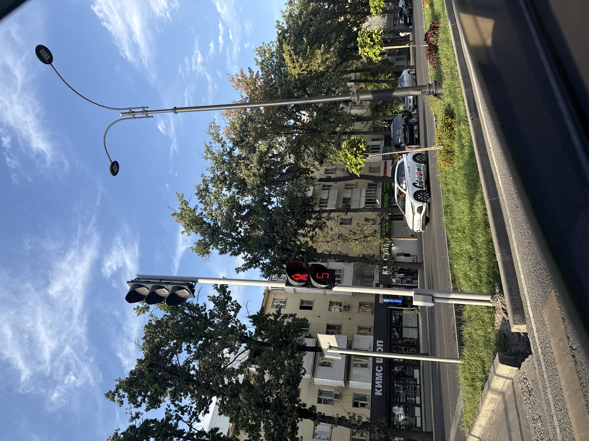 A traffic light in Bishkek, Kyrgyzstan, shows a red pedestrian signal with a countdown timer displaying "5." The traffic light is positioned on a sidewalk next to a street lined with trees and buildings. A white car with colorful branding is stopped at the intersection, indicating the pedestrian signal is active. The sky is blue with scattered clouds, and the sun casts a warm light on the scene, suggesting it is late afternoon. The buildings appear to be a mix of residential and commercial establishments, with signs visible in Cyrillic. The street is paved, and there is a grassy median between the road and the sidewalk.