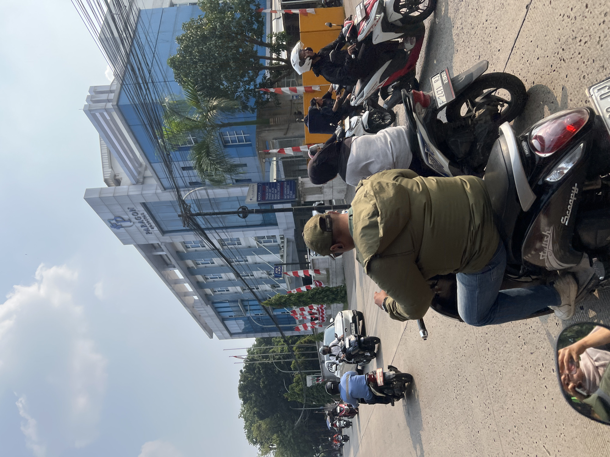 The image depicts a street scene in Citeureup, Indonesia, with a prominent building labeled "PARAGON" in the background. The street is filled with motorcycles, suggesting a busy urban environment. People are seen riding various types of motorcycles, some with passengers, navigating the road. The architecture of the Paragon building features a modern design with blue glass and white accents. Red and white flags are displayed along the building's facade, possibly indicating a national celebration or event. The overall atmosphere appears to be daytime with clear skies.