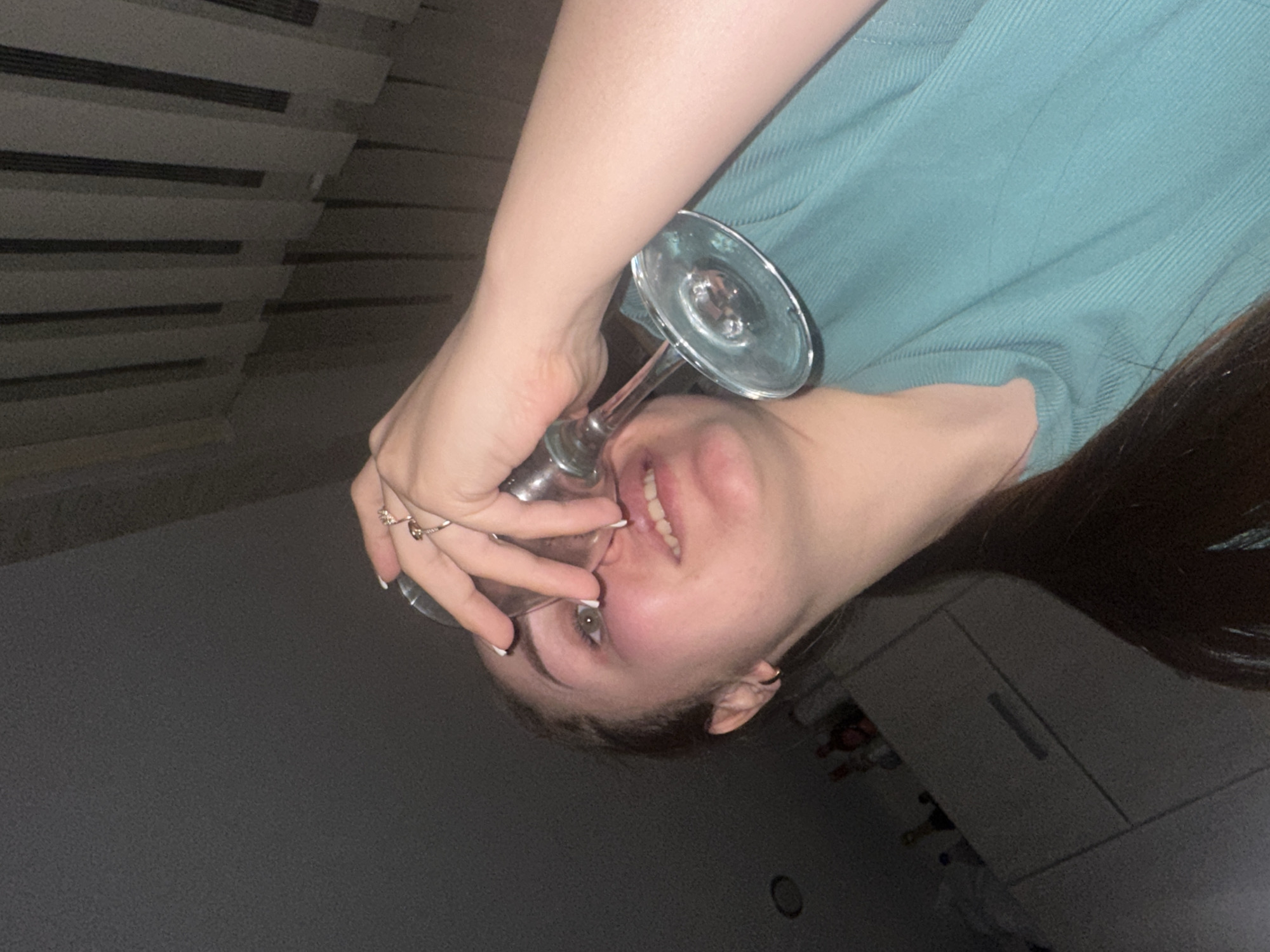 A young woman is holding a wine glass upside down against her mouth. She is wearing a light blue shirt. She has dark hair and fair skin. She is wearing rings on her fingers. The background is blurry, but appears to be a room with a window and a dresser. The location and city cannot be determined from the image.