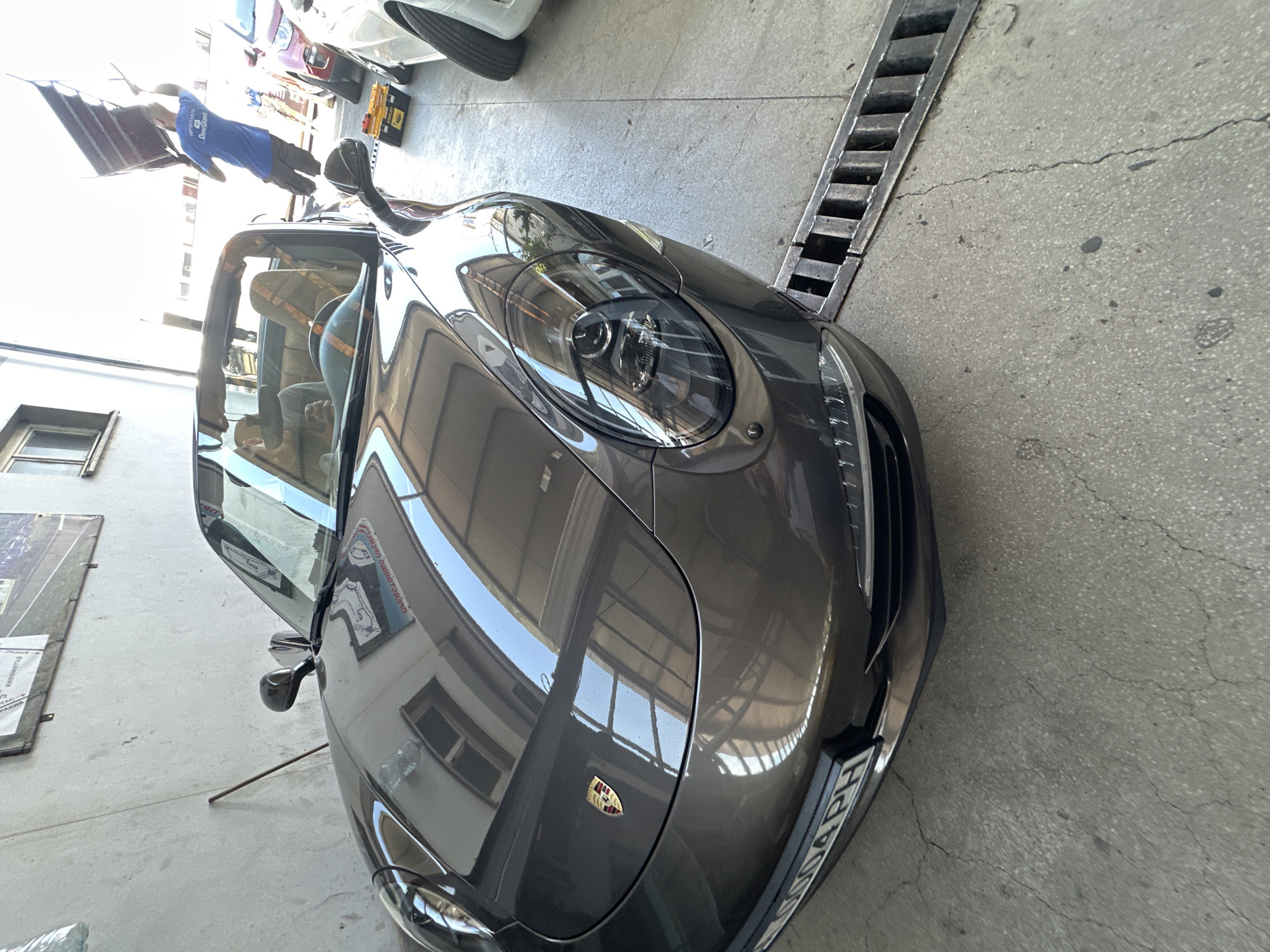A high-angle, vertical shot shows a dark gray Porsche convertible parked on a concrete floor with a drainage grate to its right. The car's front end is angled towards the right of the frame, with its headlights and emblem clearly visible. The car's interior, including the seats and dashboard, is partially visible through the open driver's side door and windshield. Sunlight reflects off the car's polished surface, indicating it is daytime.
In the background, other vehicles are visible, including a white car and a red car. A person wearing a blue shirt and dark pants is visible to the left of the car, seemingly interacting with a canopy or awning. There is also a yellow toolbox on the ground behind the person. The overall setting appears to be an indoor garage or workshop. The concrete floor shows some cracks, and a metal grate runs along the right side of the frame.
A license plate is visible on the rear of the car, with partial text reading "H004 PH." A small logo, likely the Porsche crest, is visible on the car's hood. A large banner with some Cyrillic text is also visible on the left side of the frame. The lighting suggests it is a bright, sunny day.