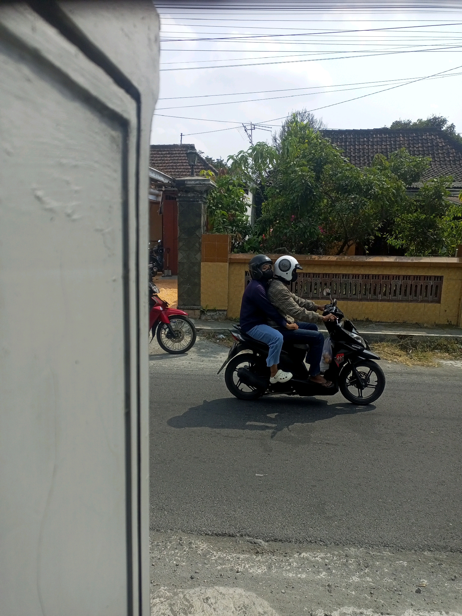 A side view captures two individuals riding a black Honda scooter on a paved road in Ngasem, Indonesia. The scooter has a black frame, black wheels, and a silver exhaust pipe. The front rider, wearing a plaid shirt and a white helmet, is holding the handlebars. The rear rider, wearing a dark purple jacket and a black helmet, has their arms around the front rider. Both individuals appear to be wearing blue jeans. The scooter's tires are casting shadows on the asphalt road. The background shows a residential area with houses, a tiled roof, a yellow wall with a wooden fence, and lush green trees. Overhead power lines are visible against a bright sky. To the left, a portion of a white building or structure is in the foreground, partially obscuring the view. A red motorcycle is parked on the side of the road in the mid-ground.