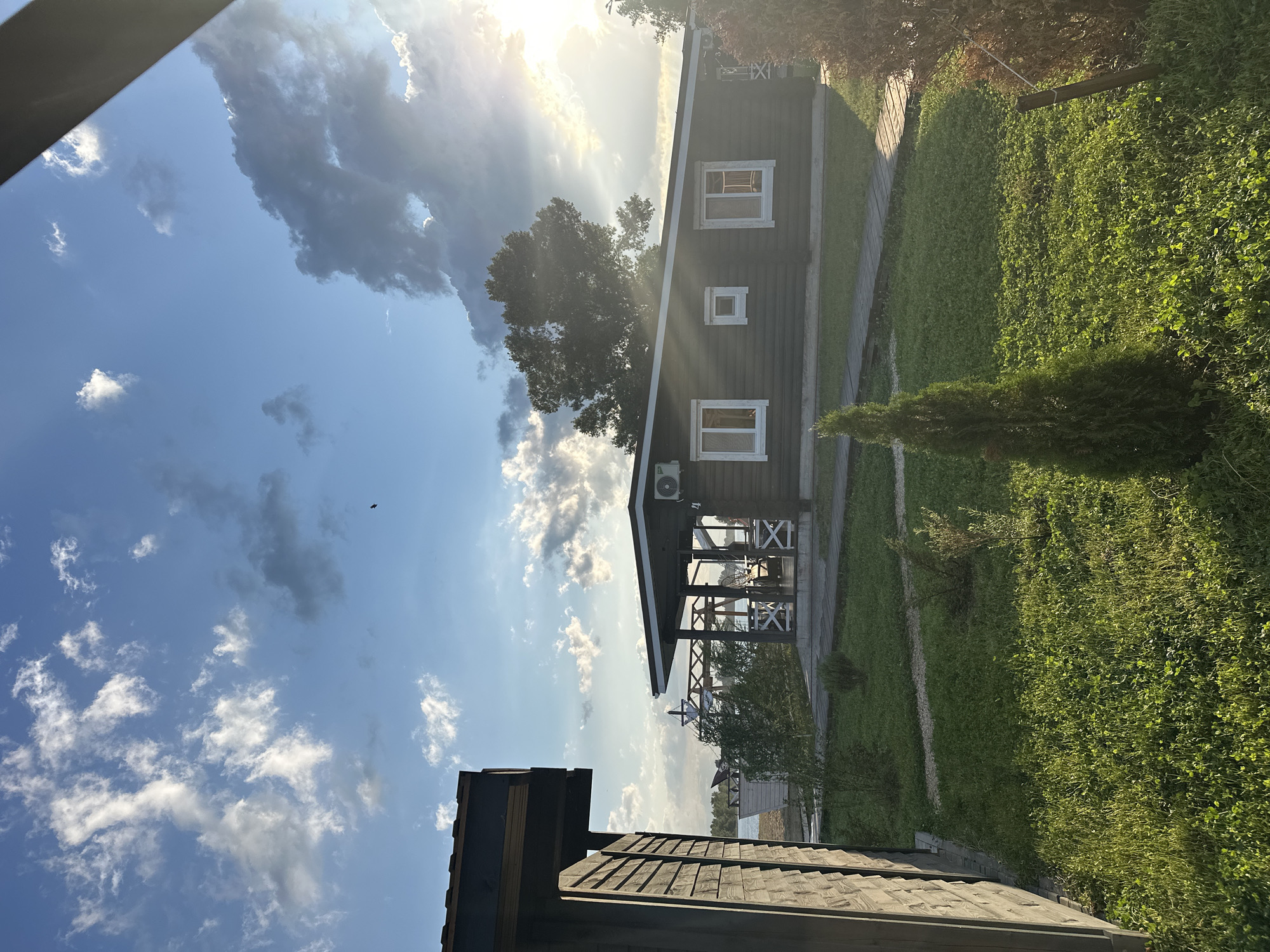 The image captures a serene outdoor scene at a waterfront property, likely a resort or private residence, under a partly cloudy sky with the sun setting. In the foreground, to the left, is the side of a wooden structure, possibly a shed or part of a building, with a dark brown wooden exterior. To the right of this structure, a dark wooden house with white-trimmed windows and a dark roof is visible. An air conditioning unit is mounted on the wall of the house. A covered porch with white lattice railings extends from the front of the house, furnished with outdoor seating.
The house is situated on a well-maintained lawn with scattered evergreen bushes and a small, conical evergreen tree in the middle ground. A gravel path leads from the house to what appears to be a wooden walkway or pier leading towards a body of water, suggested by the glint of sunlight on its surface and a distant view of a sandy shore with a white structure. Tall trees form a backdrop to the house, and the sun's rays are piercing through the clouds, creating a dramatic effect.
The overall impression is one of peacefulness and natural beauty, with the architecture blending harmoniously with the environment. While no people are explicitly visible, the presence of outdoor furniture suggests recent or anticipated human activity. The location in Balakovo, Russia, would place this scene likely along the Volga River, a prominent feature of the city.