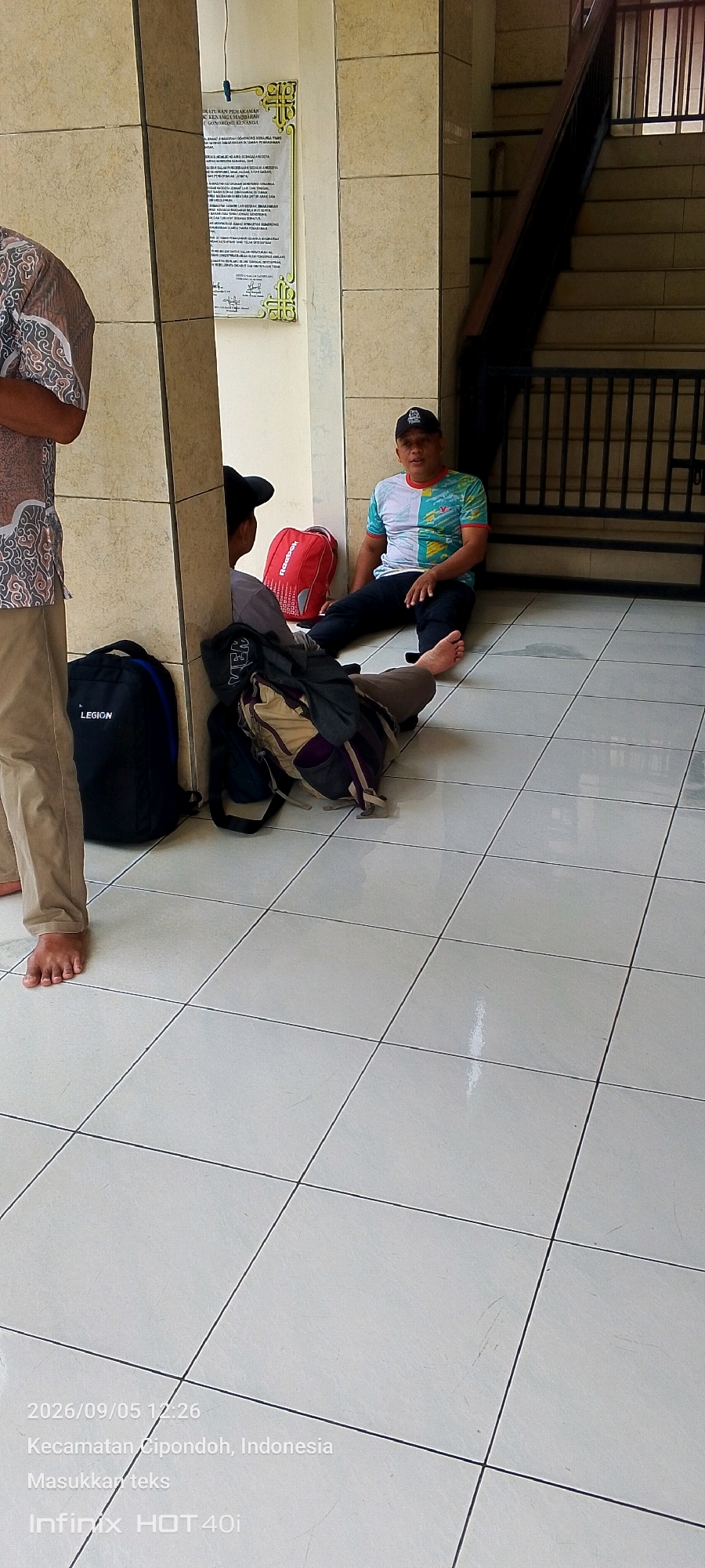 Two men are seated on the tiled floor near a pillar and a staircase. The man on the left is partially visible, wearing a patterned shirt and khaki pants. He is barefoot. The man on the right wears a colorful t-shirt and dark pants and is also barefoot. He is looking towards the camera and speaking. In the foreground, several backpacks are on the floor: a black "LEGION" backpack, a red backpack, and another backpack with clothing piled on top of it. A notice is posted on the pillar behind the men. The environment appears to be an indoor public space, possibly a community center or building lobby, given the presence of the notice and the tiled flooring. The timestamp on the image indicates the location as Cipondoh, Indonesia, within the city of Tangerang.