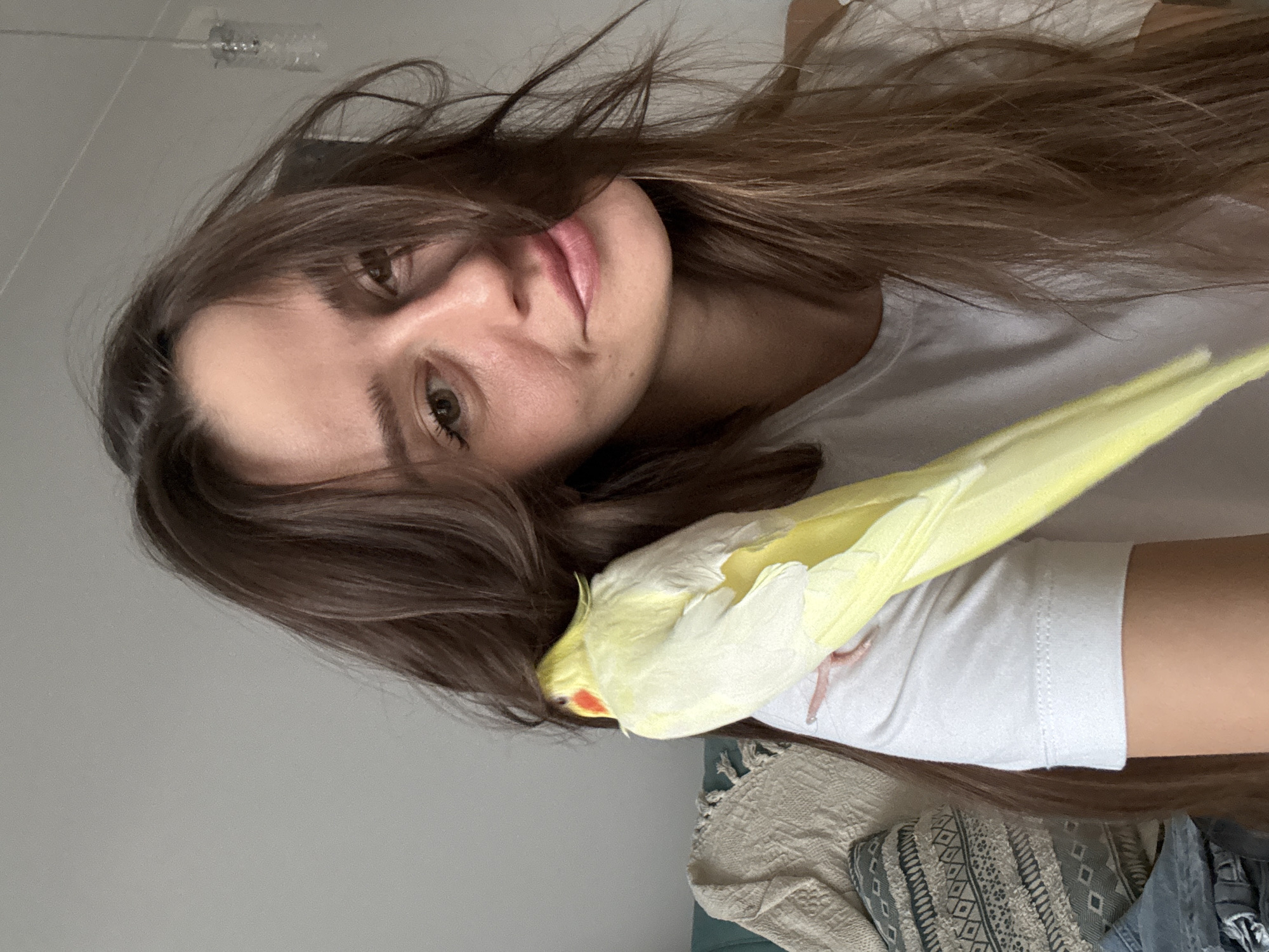 A light-skinned woman with long brown hair is holding a yellow and white cockatiel. The bird is perched on her arm. She is wearing a white t-shirt. The background is a light gray wall. The photograph does not show any evidence of the location being in the city of Krasnoyarsk, Russia.