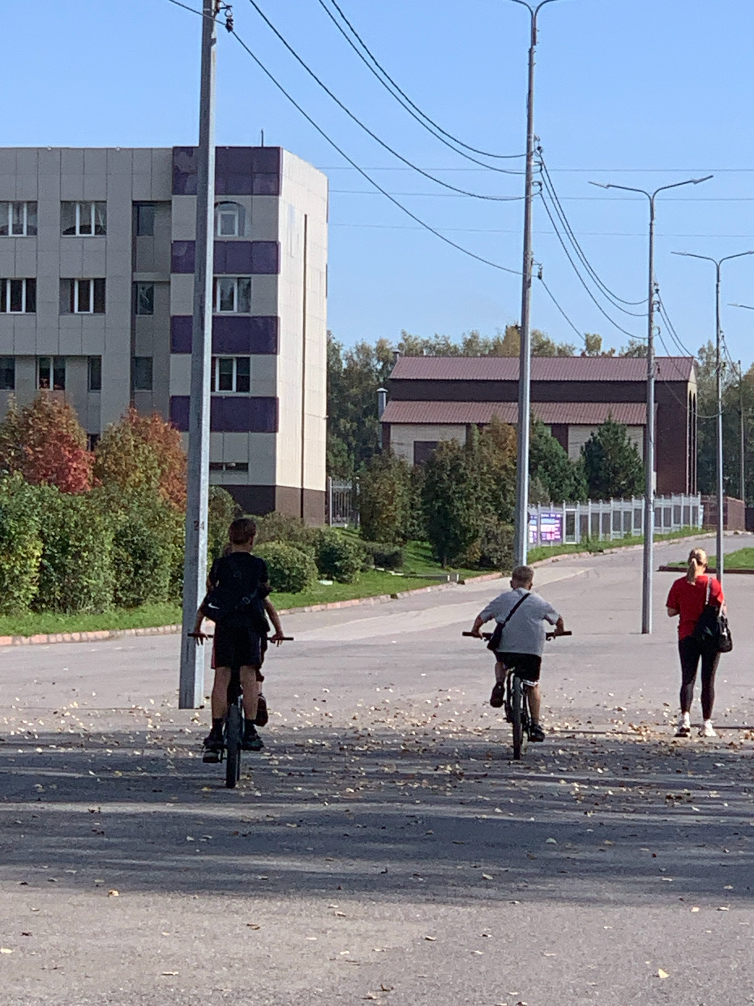 The image depicts an outdoor scene with a clear blue sky. In the foreground, a paved road is covered with fallen leaves. Three people are visible. On the left, a person wearing a black t-shirt and shorts is riding a bicycle away from the camera. In the center, a person in a grey t-shirt and dark shorts is also riding a bicycle away from the camera. On the right, a woman in a red t-shirt and dark leggings is walking away from the camera, carrying a black bag over her shoulder. In the background, a tall, multi-story building with a grey facade and purple accents stands on the left. To the right of this building, there are trees with autumn foliage. Further in the background, a long, brown-roofed building is visible, and a white fence runs along the side of the road. Several lampposts are positioned along the road, with power lines running between them. A thin, vertical pole with a number "24" is also present. The scene appears to be a street in Prokopyevsk, Russia, likely during the autumn season given the leaves on the ground and trees.