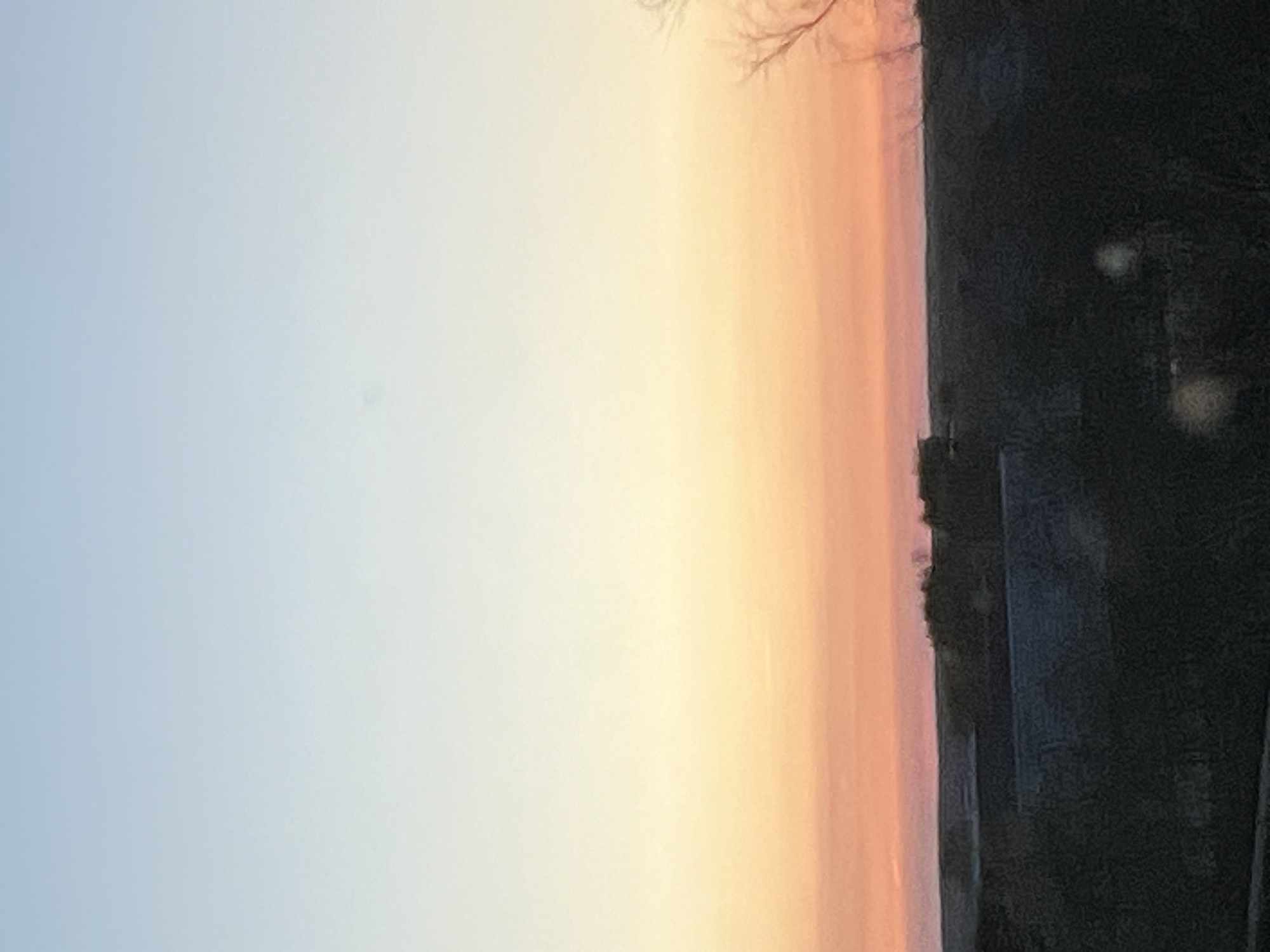 The image depicts a landscape at sunset or sunrise, with a clear gradient in the sky from light blue to yellow and orange. On the right side, there is a dark mass, possibly trees and buildings. It is difficult to identify the exact location as it lacks distinctive landmarks.