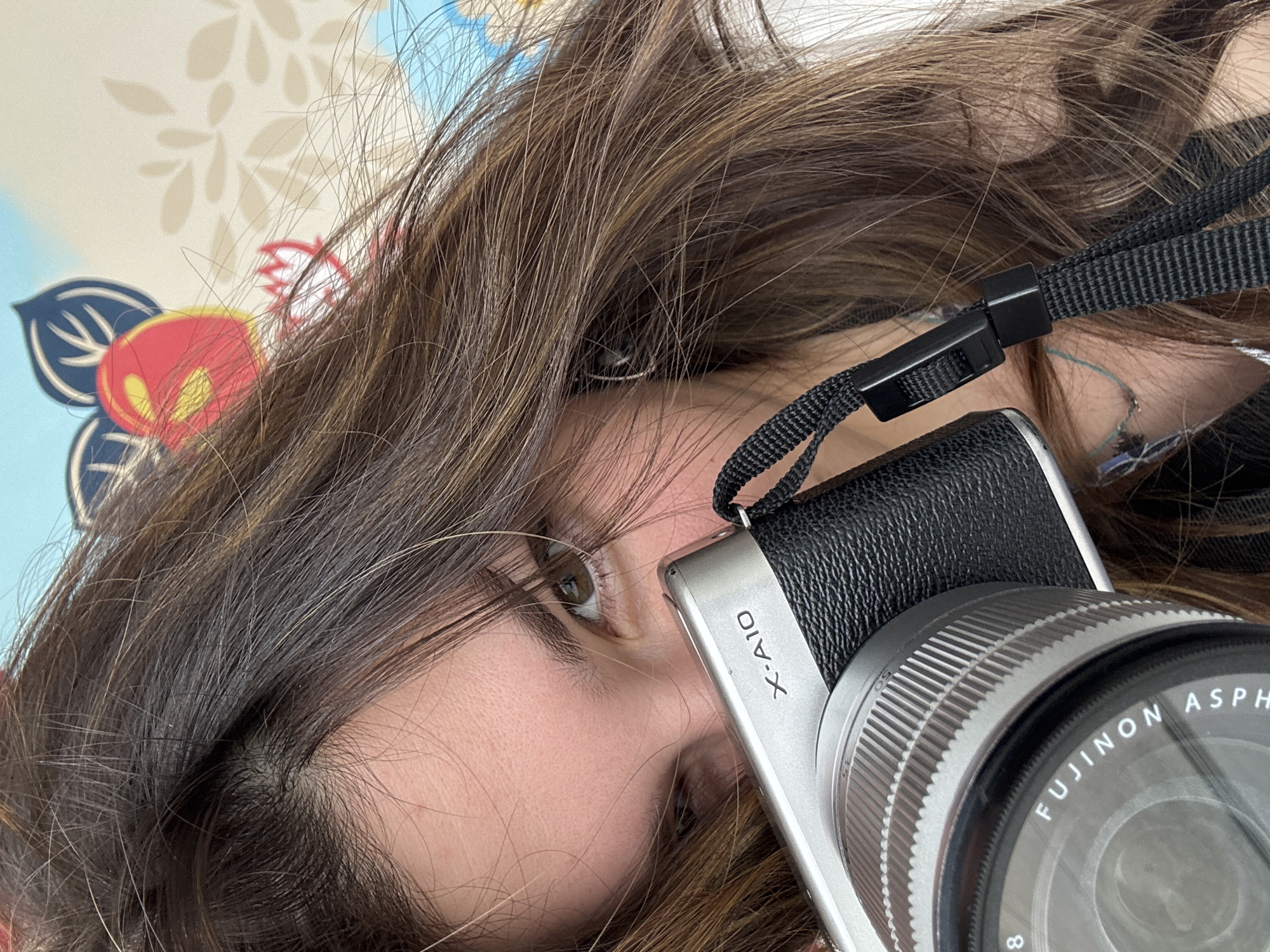 A close-up, angled shot features a person, likely female, with a camera. The subject's face is partially visible, showing one brown eye, part of the nose, and cheek, framed by abundant long, wavy brown hair, which includes some lighter strands spread across the image. The person appears to be looking directly at the viewer with a neutral expression.
A silver Fujifilm X-A10 mirrorless camera with a black textured grip is prominently positioned in the lower right and center of the frame, held close to the person's face. Visible text on the camera body clearly identifies it as "X-A10". The attached lens is a silver Fujinon lens, with the text "FUJINON AS..." (likely ASPH) and the number "8" visible on its barrel. A black camera strap with a plastic quick-release buckle is also visible, connected to the camera body.
The background consists of a patterned surface, likely part of bedding such as a pillowcase or duvet cover, featuring abstract floral and leaf motifs in shades of blue, light yellow, red, and off-white/beige. The lighting suggests an indoor setting, possibly during the daytime or a well-lit evening. While the contextual location is Kanazawa, Japan, there are no specific visual cues within the image itself to denote this geographical location.