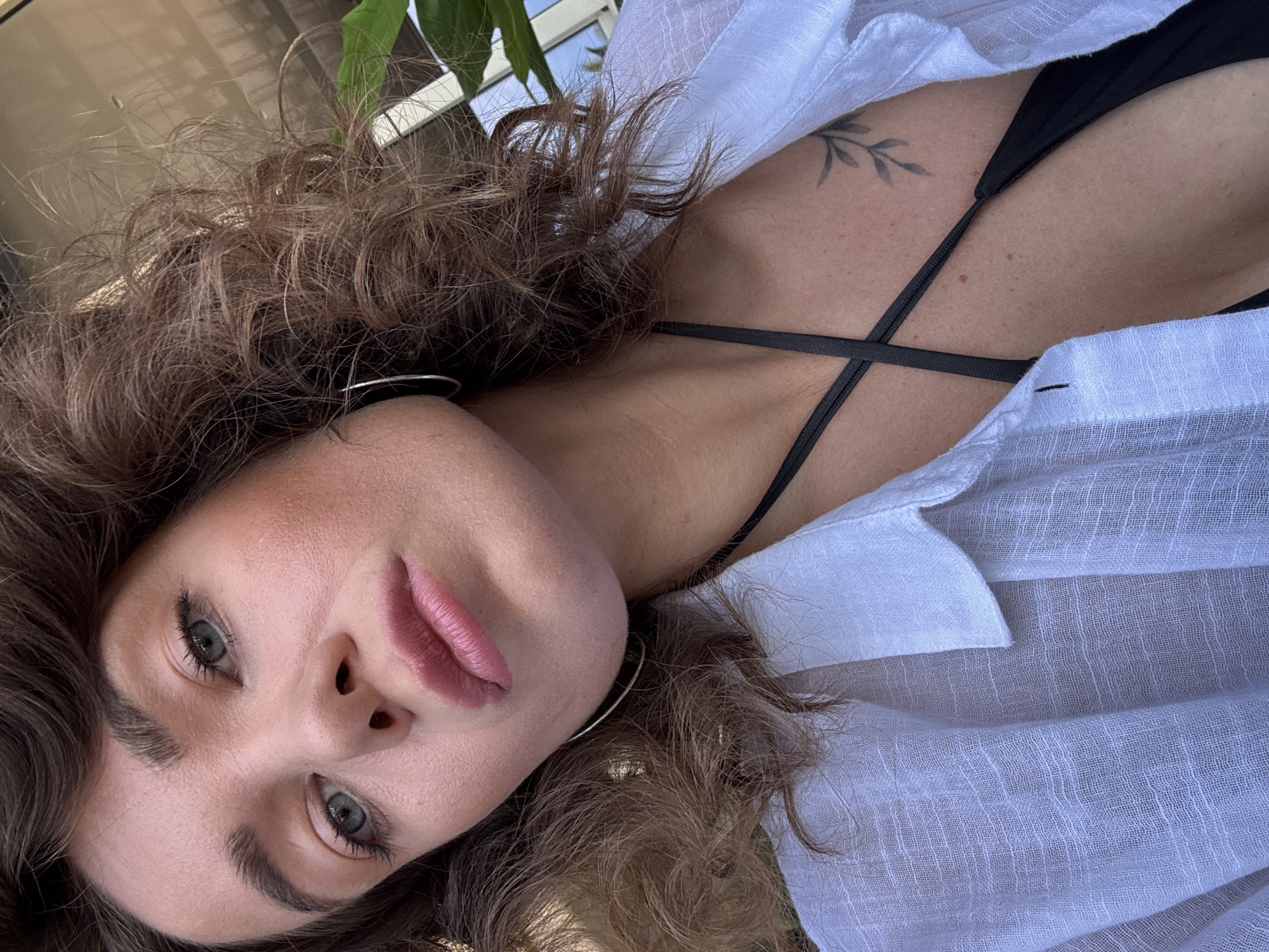 A close-up, tilted portrait captures a person, likely female, from a high-angle perspective. Her head is angled almost horizontally within the frame, with her gaze directed straight at the viewer. She has curly, brown hair that frames her face. Her eyes are a striking green/hazel, and she wears natural-looking makeup, including defined eyebrows, mascara, and pink lipstick. Silver hoop earrings are visible on both ears.
She is dressed in a white, sheer, textured button-up shirt, unbuttoned to reveal a black bikini top or bra with thin, criss-cross straps. Her shoulder and upper chest are visible, showing fair skin with a few moles or freckles. A small, stylized tattoo of a leafy branch is etched onto her right shoulder/collarbone area.
The background is softly blurred, showing hints of green foliage and what appears to be reflections of a window or building, suggesting an outdoor or semi-outdoor setting. The lighting is bright and natural, indicating daytime. The overall mood conveyed is calm and direct. There is no visible text in the image. The location context for this scene is Prijevor, Montenegro.