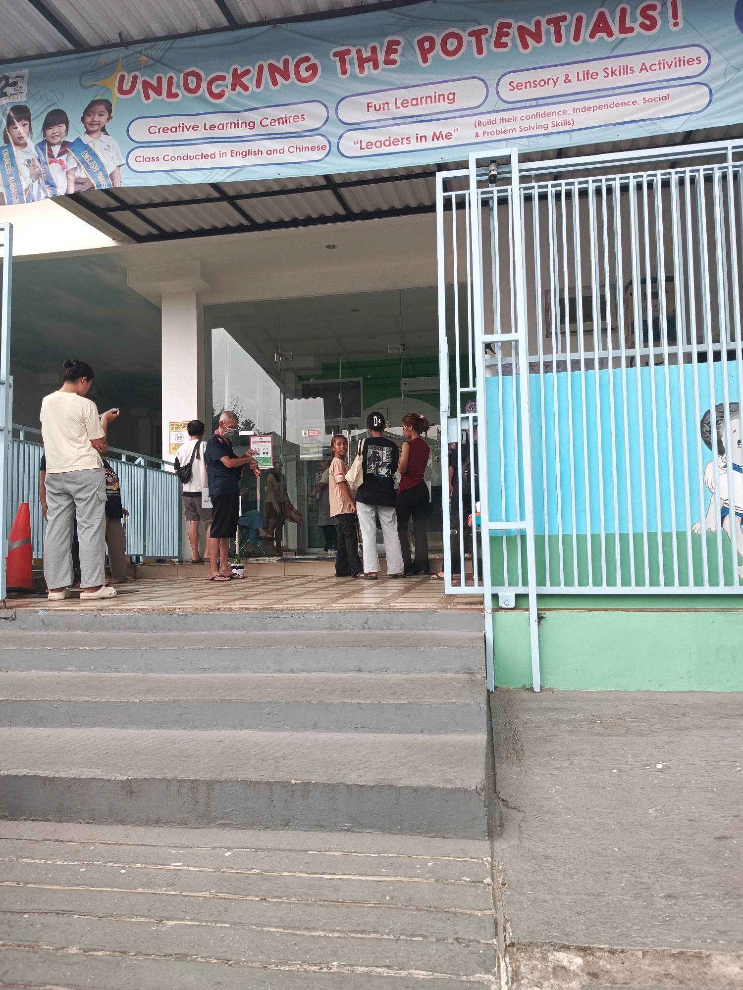 The image depicts the entrance to a learning center in Pakulonan Barat, Indonesia. A banner above the entrance reads "UNLOCKING THE POTENTIALS!" and lists various programs such as "Creative Learning Centres," "Fun Learning," and "Sensory & Life Skills Activities." Several individuals are visible standing near the entrance, some waiting and others engaged in conversation. The environment includes a white gate, a blue and green painted wall with a cartoon dog illustration, and concrete steps leading up to the building's entrance, which features glass doors. A red traffic cone is also present on the left side of the image.