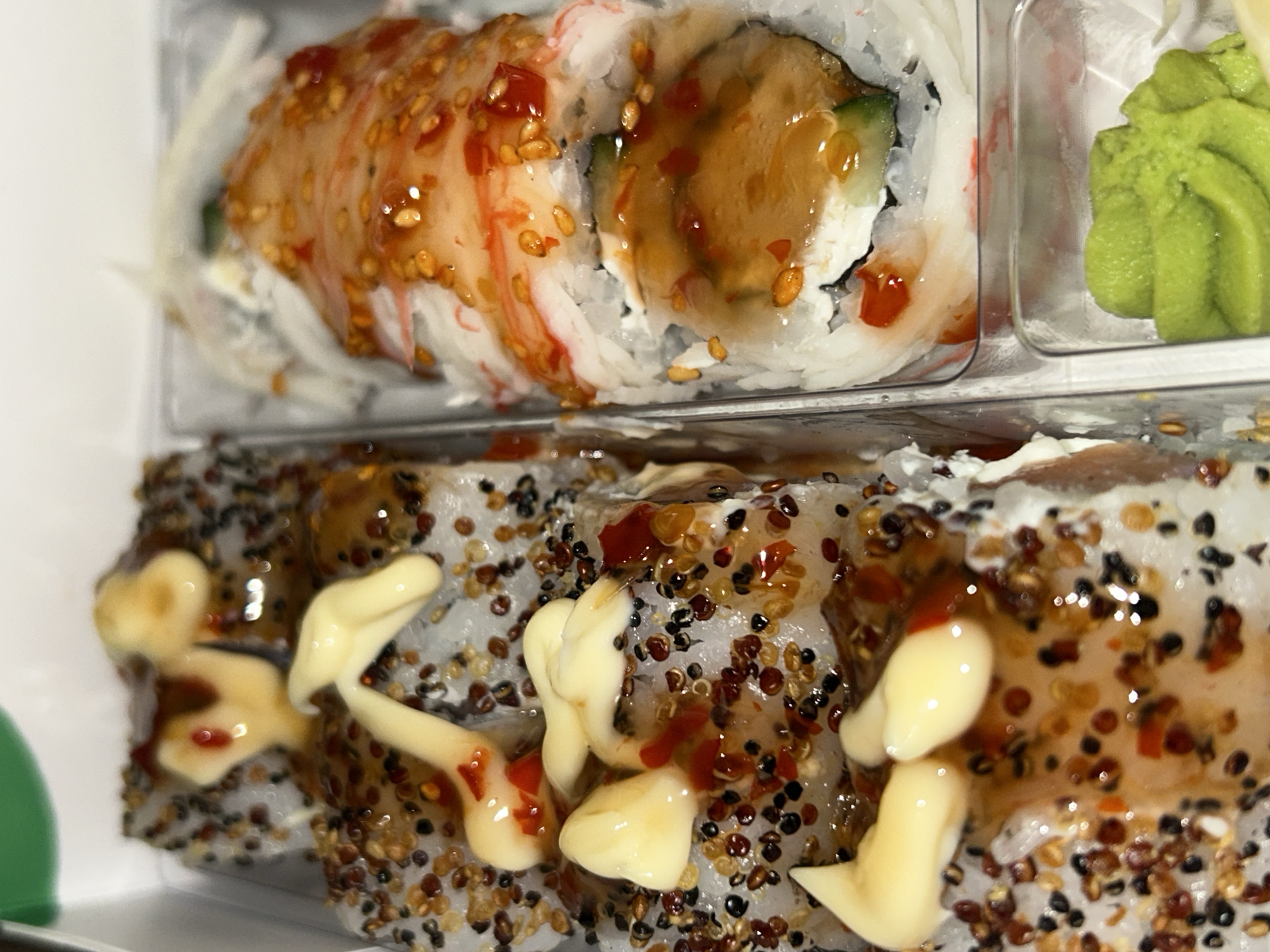 The image shows two trays of sushi rolls. The top tray contains two rolls with visible crab, cucumber, cream cheese, and a brown sauce with sesame seeds. There is also a portion of wasabi in a separate compartment. The bottom tray shows rolls covered in seeds and topped with a creamy sauce and a red sauce.
