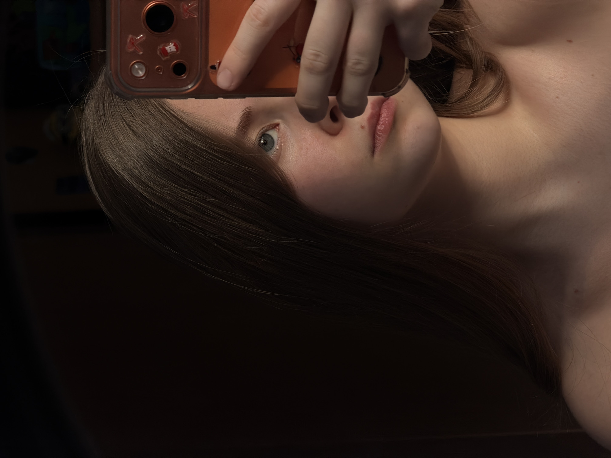 A person is holding an orange cell phone above their face, obscuring their forehead and partially their eye. The individual has light skin, light brown hair, and blue eyes. The background is mostly dark, with some indistinct objects to the left. The location is unknown and cannot be determined from the image.