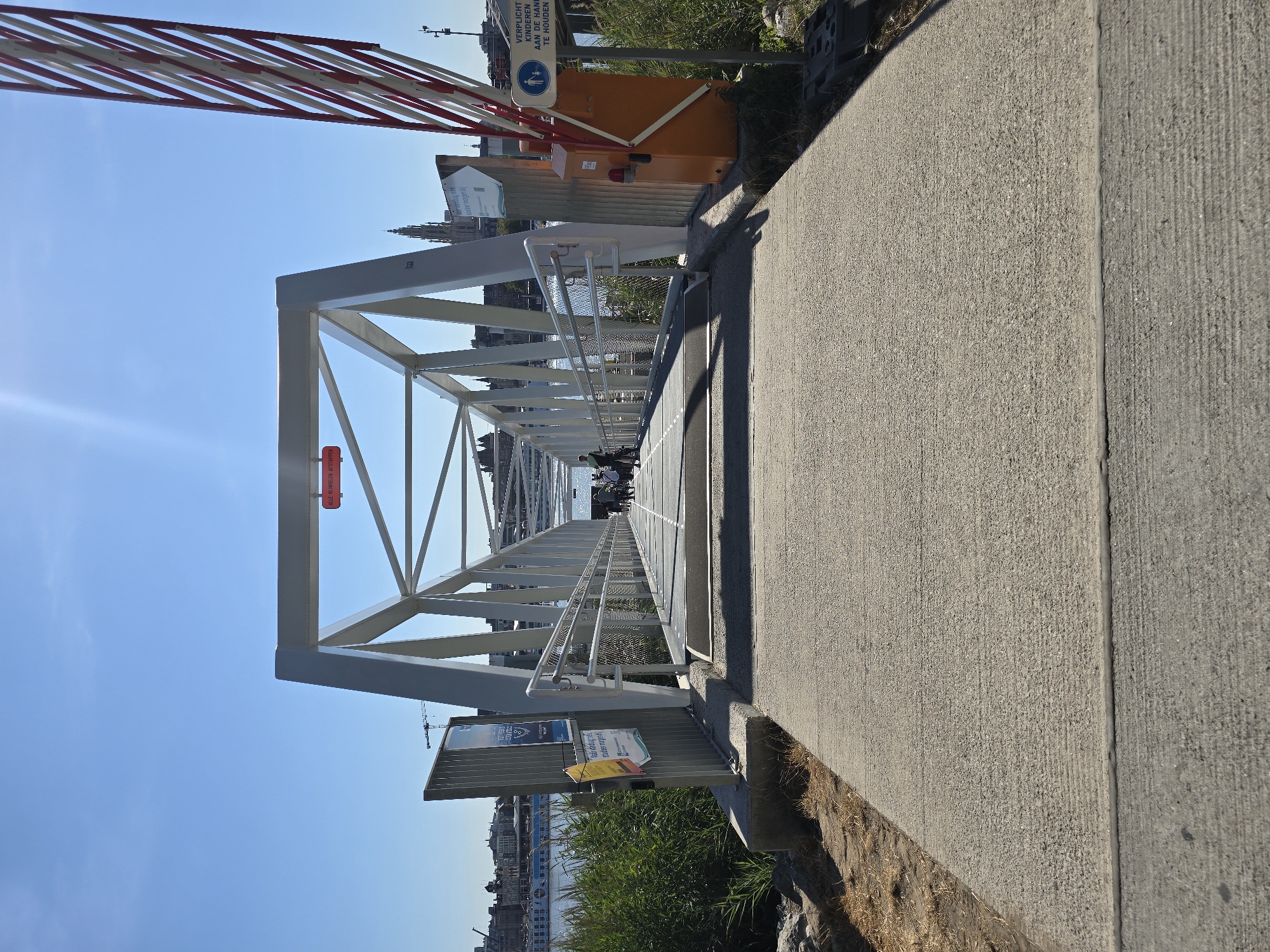 The image shows a pedestrian bridge over water in Antwerp, Belgium. The bridge is made of white metal and has a long, narrow walkway. On the right side of the bridge, there is a red and white striped barrier and an orange control box. There are signs with instructions and warnings. Several people are walking across the bridge, some with bicycles. The sky is clear and blue, and the sun is shining. In the background, there are buildings and a crane.