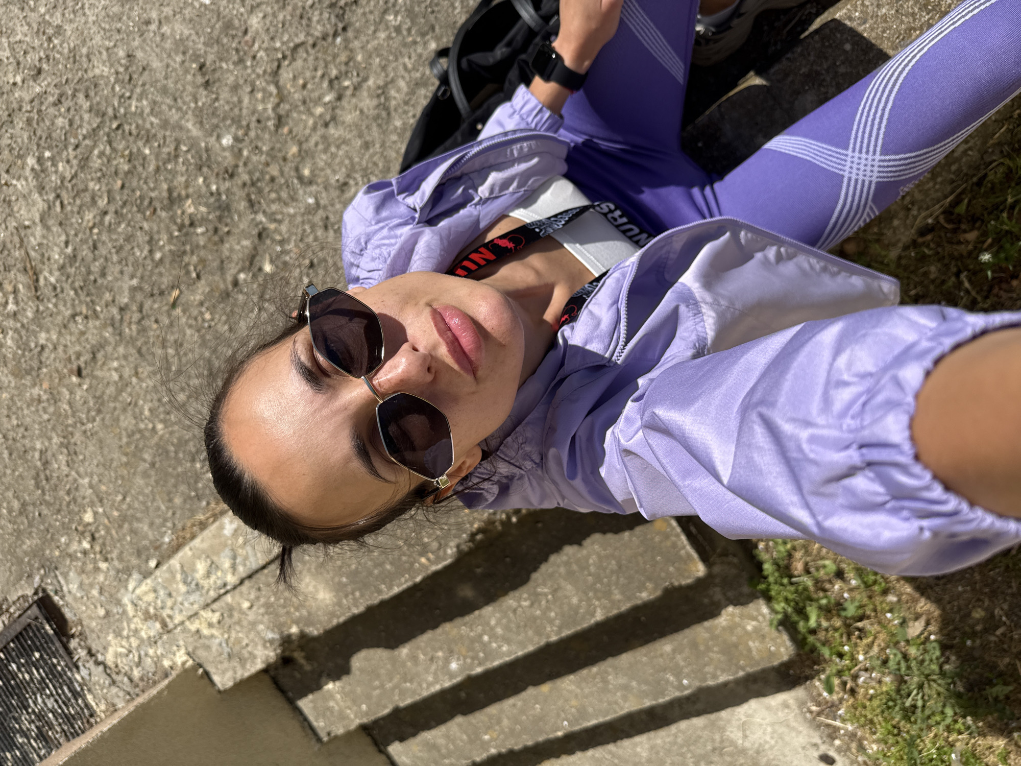 A high-angle selfie captures a young person, likely female, reclining or seated outdoors on a sunny day in Pleven, Bulgaria. The subject is looking directly up at the camera, wearing dark, hexagonal sunglasses with gold frames. Their dark hair is pulled back.
They are dressed in a casual athletic outfit consisting of a lilac or lavender windbreaker jacket and matching high-waisted leggings. The leggings feature a distinctive white grid pattern on the outer thighs. A white sports bra is visible beneath the open jacket, with a black strap that has "NURSE" written in white capital letters on it, along with a red graphical element. On their left wrist, a black smartwatch is visible. A black backpack is partially seen behind their back.
The setting appears to be an outdoor urban or semi-urban environment. The person is situated on rough, light grey concrete ground, possibly steps, as strong shadows from distinct edges are cast onto the surface, indicating bright sunlight. A dark metal drain grate is visible in the lower left corner of the frame. Patches of sparse green grass are also visible towards the right side of the image. The overall impression is one of a relaxed moment captured in direct sunlight.