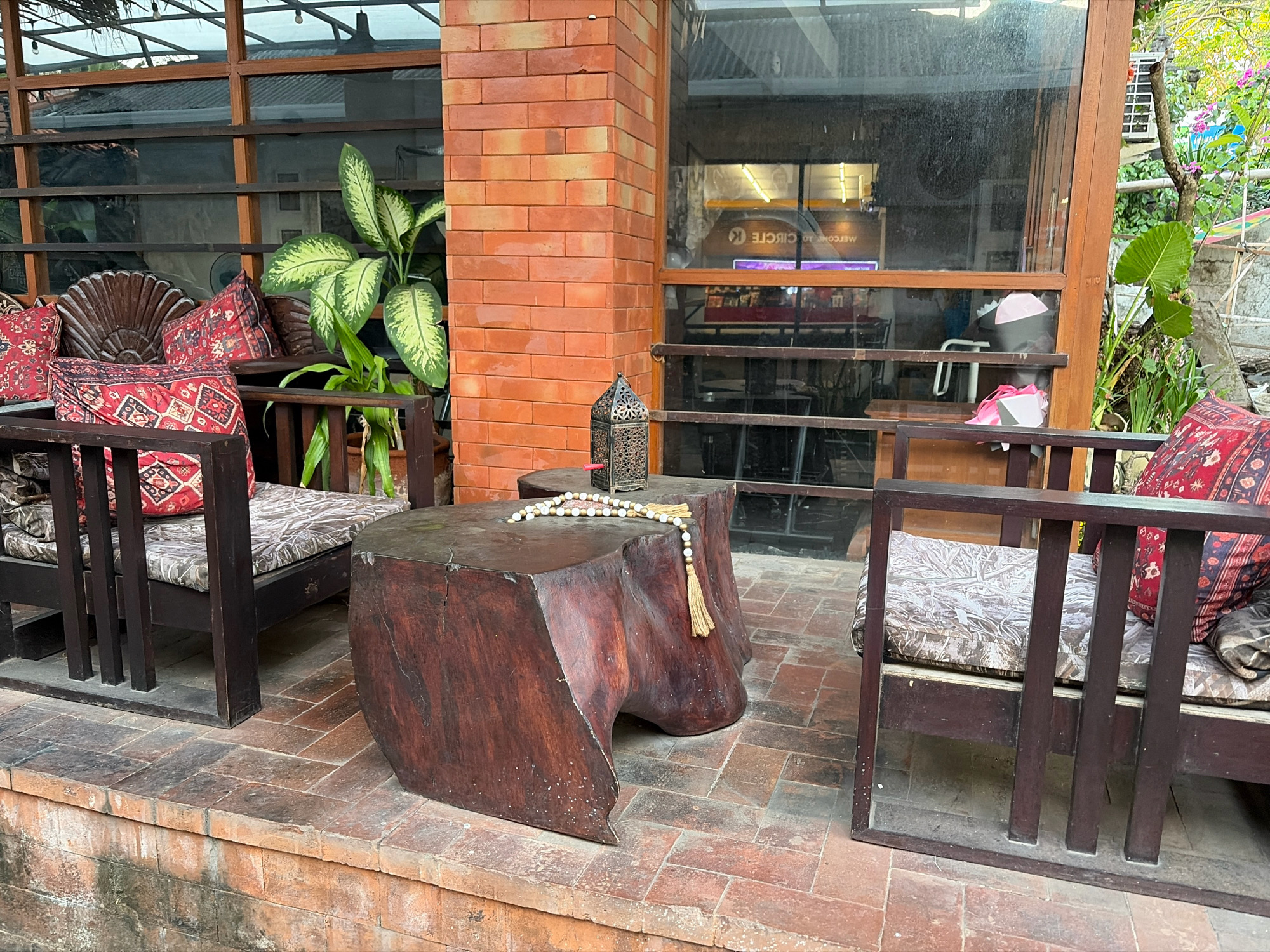 A covered outdoor seating area features two dark wooden benches with patterned cushions and red and black patterned throw pillows. A large, irregularly shaped wooden table sits between the benches, adorned with a string of beads with a tassel. On the table, a decorative metal lantern is visible. Behind the seating area is a brick wall and large windows that appear to look into an interior space. A large potted plant with broad green and white leaves is positioned to the left of the seating. The floor is paved with reddish-brown tiles. The overall setting suggests a cafe or restaurant patio in Kuta, Indonesia.